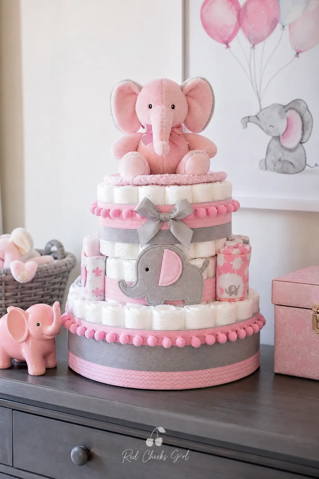 Pink Elephant Diaper Cake — Soft pink and heather grey diapers alternate by tier, with a small plush elephant seated on top and grey pom pom trim marking each