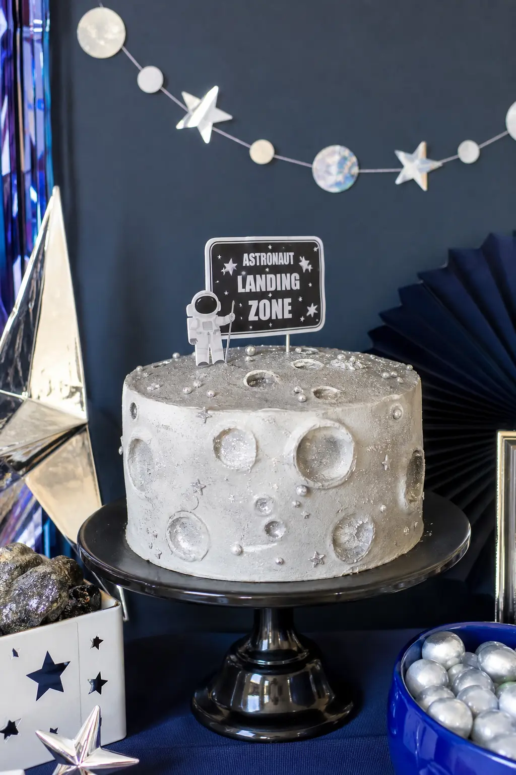 Astronaut Landing Zone Display — A textured grey and white fondant cake, styled to look like cratered moon rock, sits on a black stand for maximum contrast.
