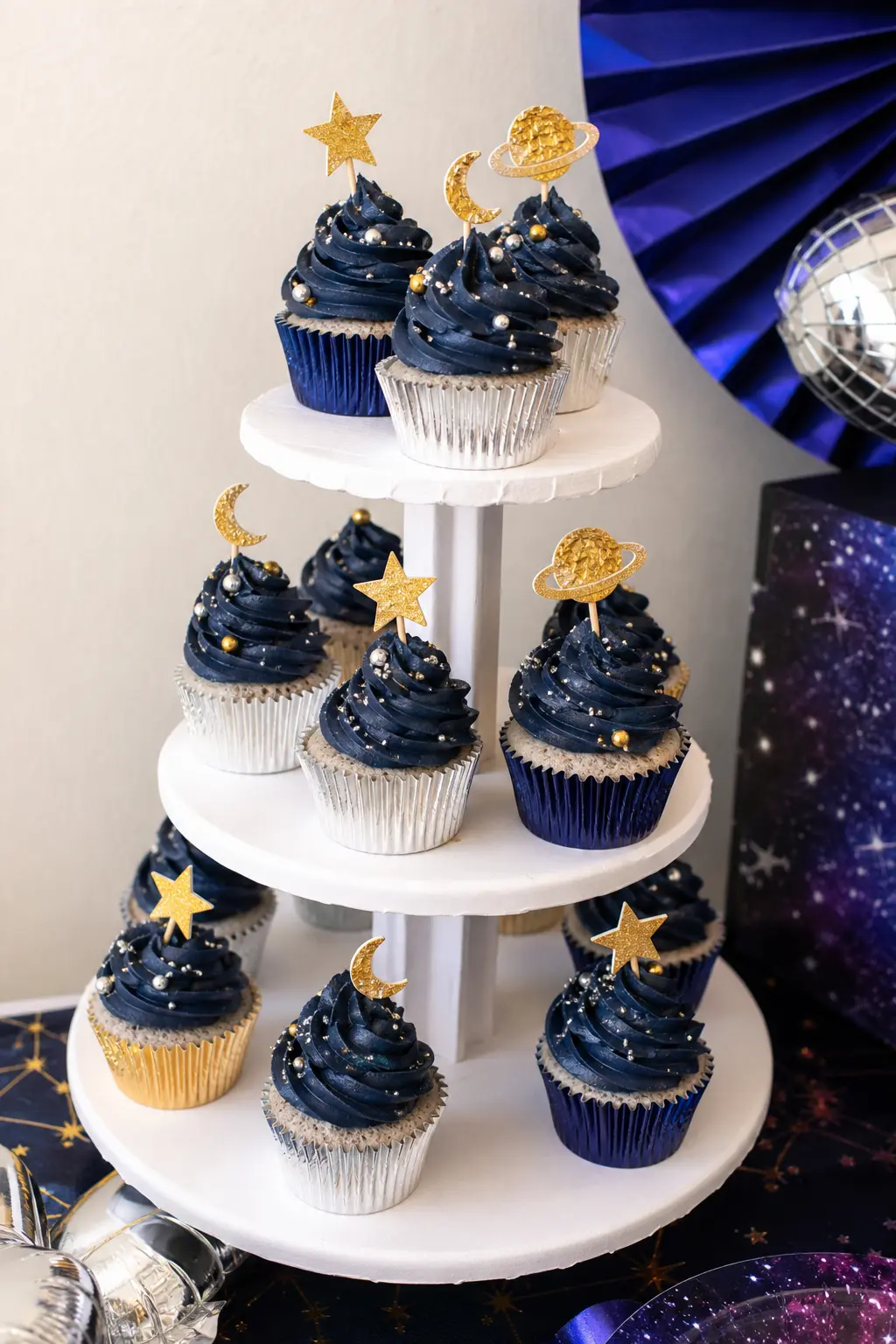 Cosmic Cupcake Tower — Navy buttercream swirls, finished with silver dragees and small gold foil toppers, arranged on a plain white tiered stand, do most of