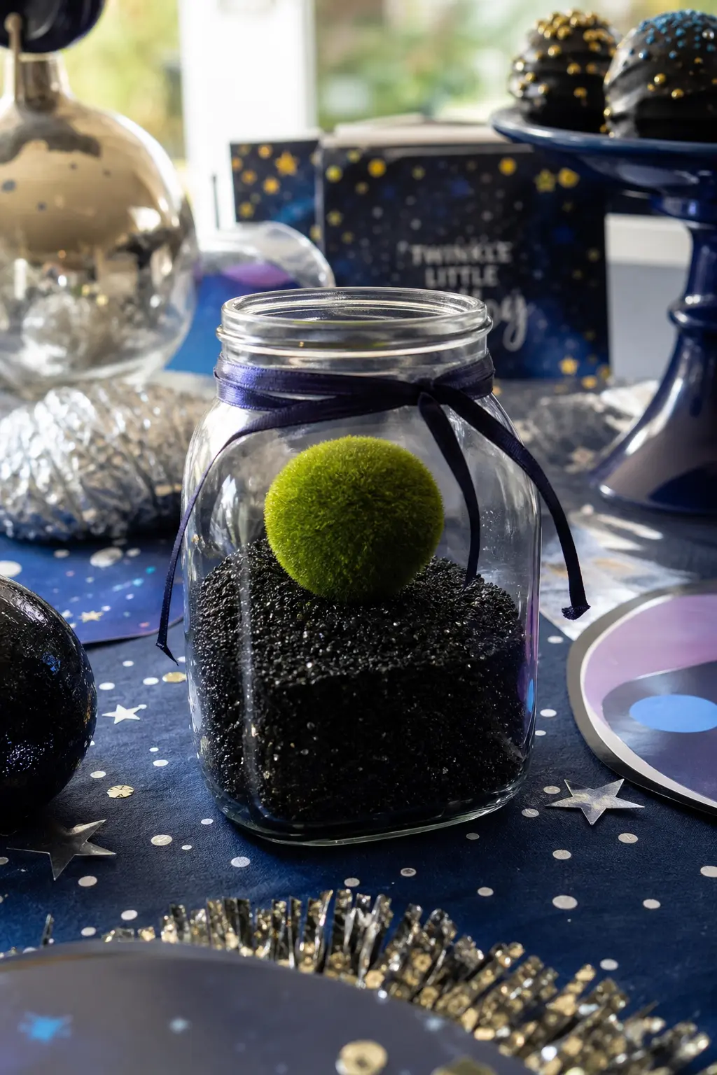 Mini Terrarium Planet Jar — A small moss green sphere set into black sand at the base of a clear glass jar reads as a tiny planet under glass rather than a