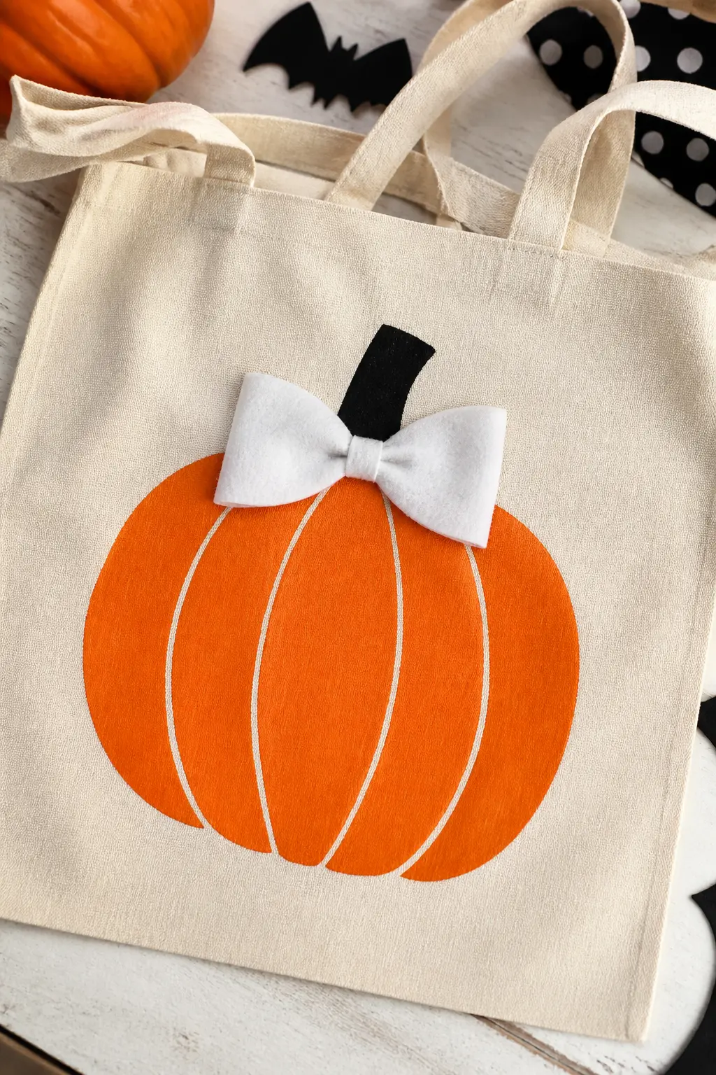 Felt Bow on a Painted Pumpkin — Paint or stamp an orange pumpkin on the bag front, then cut a bow from white felt: two rectangles pinched at the centre and a