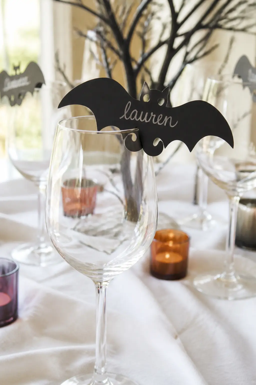 Paper Bat Name Tag — Cut bat shapes from black card stock, notch the wing to slot over the rim of a wine glass, and write a guest's name in a silver or grey