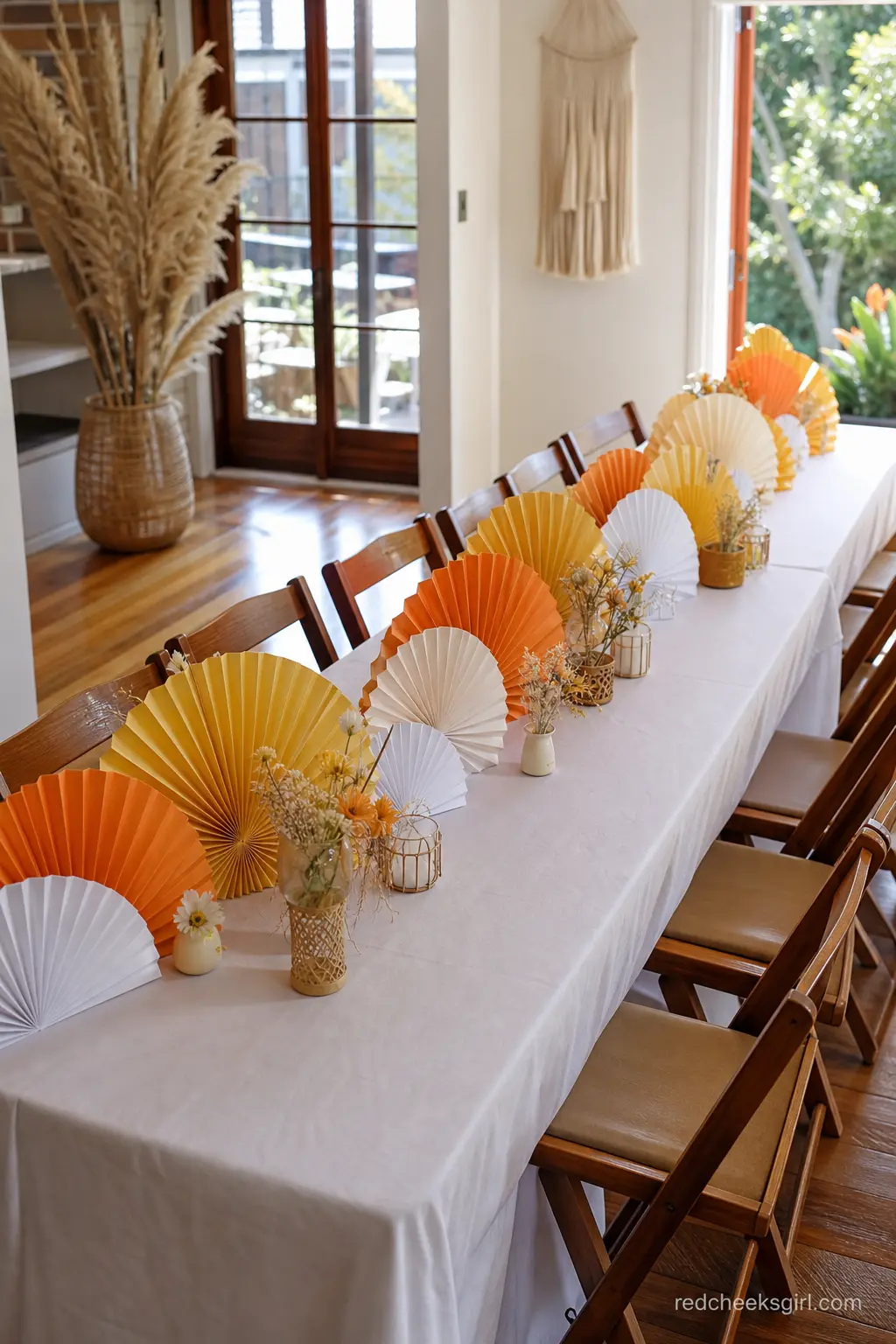 Long Communal Table with Paper Fan Sunbursts — Layered paper fans in yellow, orange and white, arranged like a row of overlapping suns, make a striking