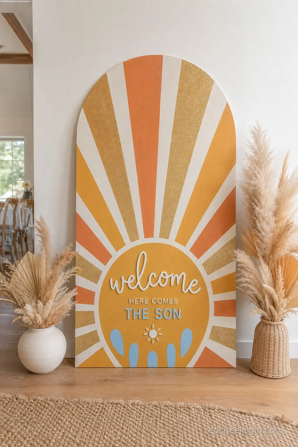 Personalized Sunburst Welcome Sign — A wooden arch sign painted with radiating sunburst rays in orange and gold, leaning against a wall near the gift table