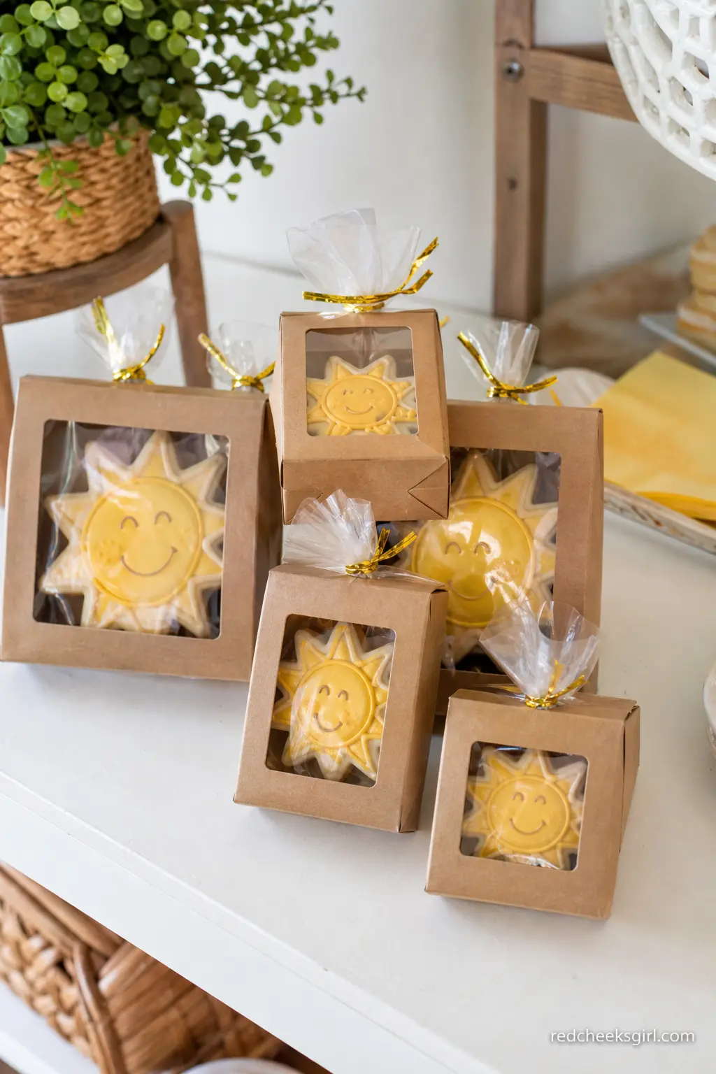 Sun Cookie Favor Boxes — Small kraft boxes holding a single sun shaped cookie, tied with a thin gold twist tie, stack neatly on a shelf near the exit and give