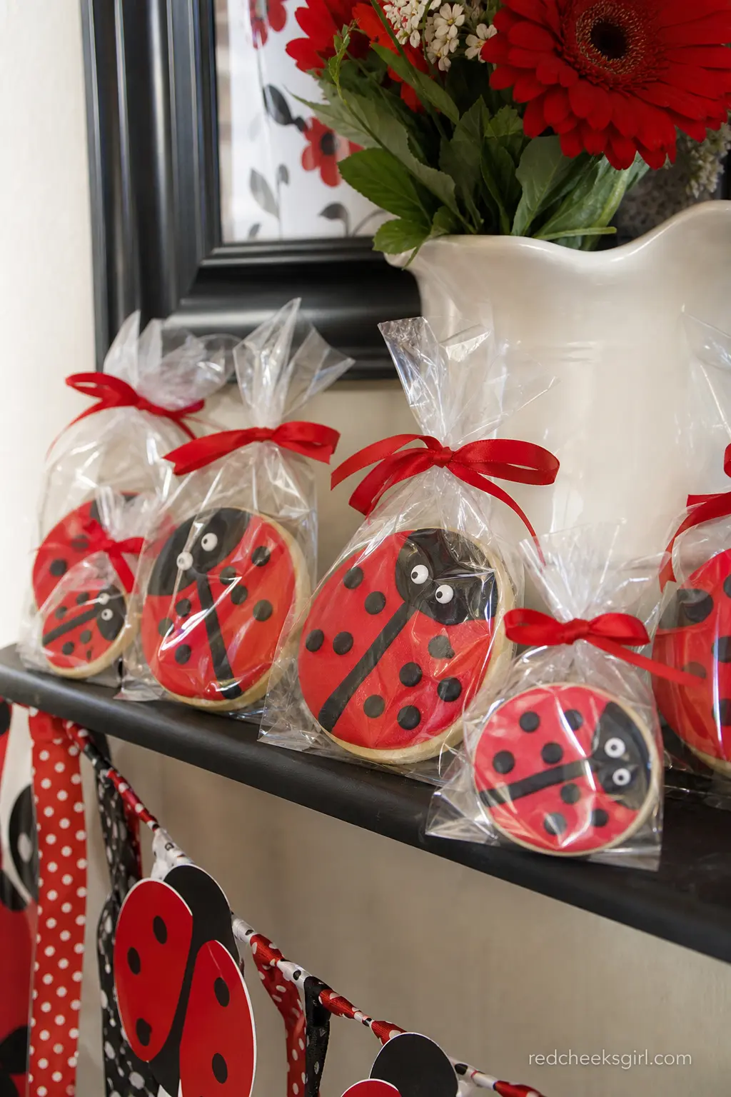 Ladybug Cookie Favor Box Display — Iced sugar cookies shaped like ladybugs in red, black and white sit stacked inside clear favor boxes tied with red satin