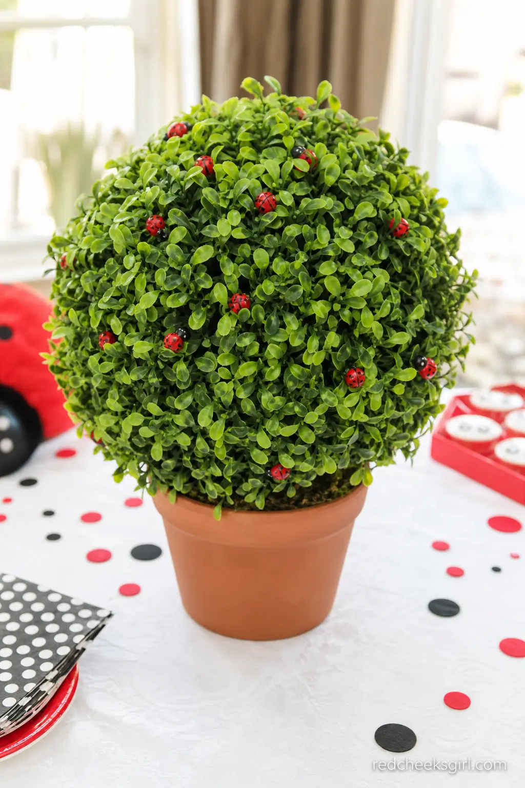 Ladybug Topiary Centerpiece — A single faux boxwood topiary sits in a terra cotta pot on a white tablecloth, with small red felt ladybugs clipped among the