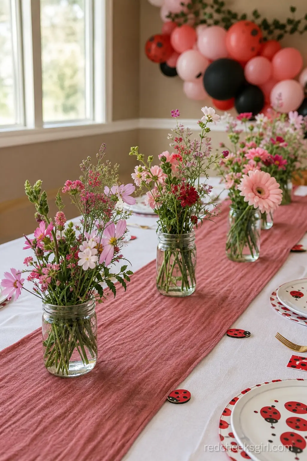 Wildflower Jam Jar Bouquet Runner — Small mismatched jam jars filled with pink and red wildflowers march down a dusty rose table runner, spaced so the table