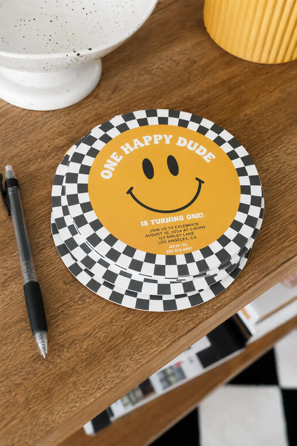 Retro Round Invitation Display — A small stack of invitations with a black and white checker border and a mustard smiley centered on the front, left on the