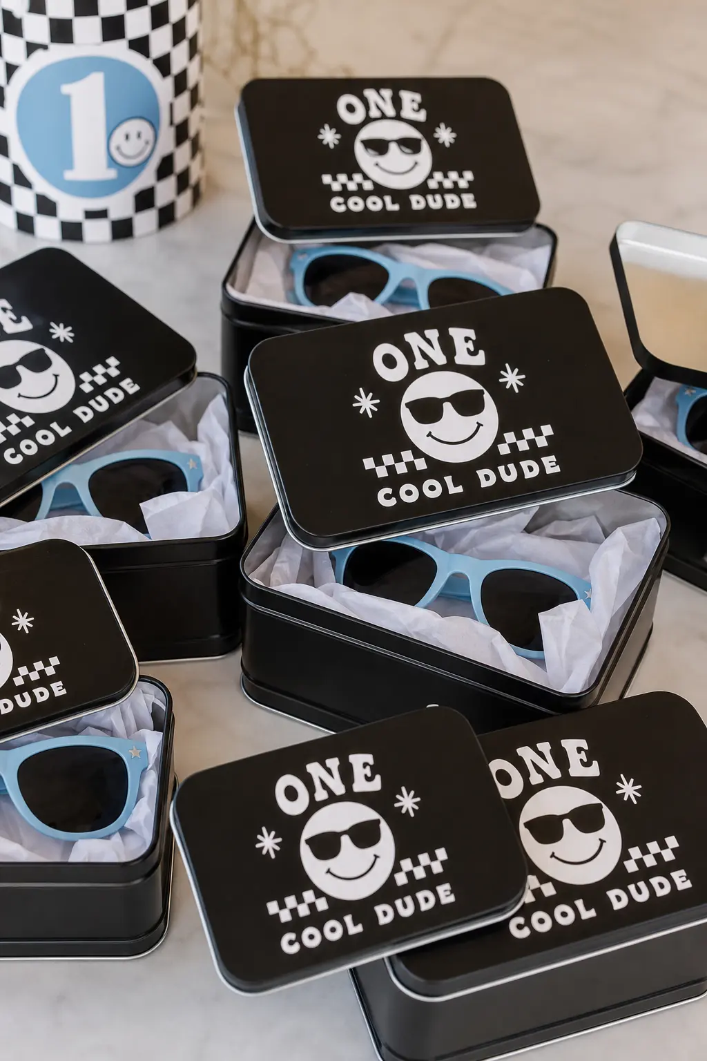 Retro Sunglasses Favor Tins — A cluster of black metal tins, each holding a pair of sky blue kids' sunglasses wrapped in white tissue, works as both a favor