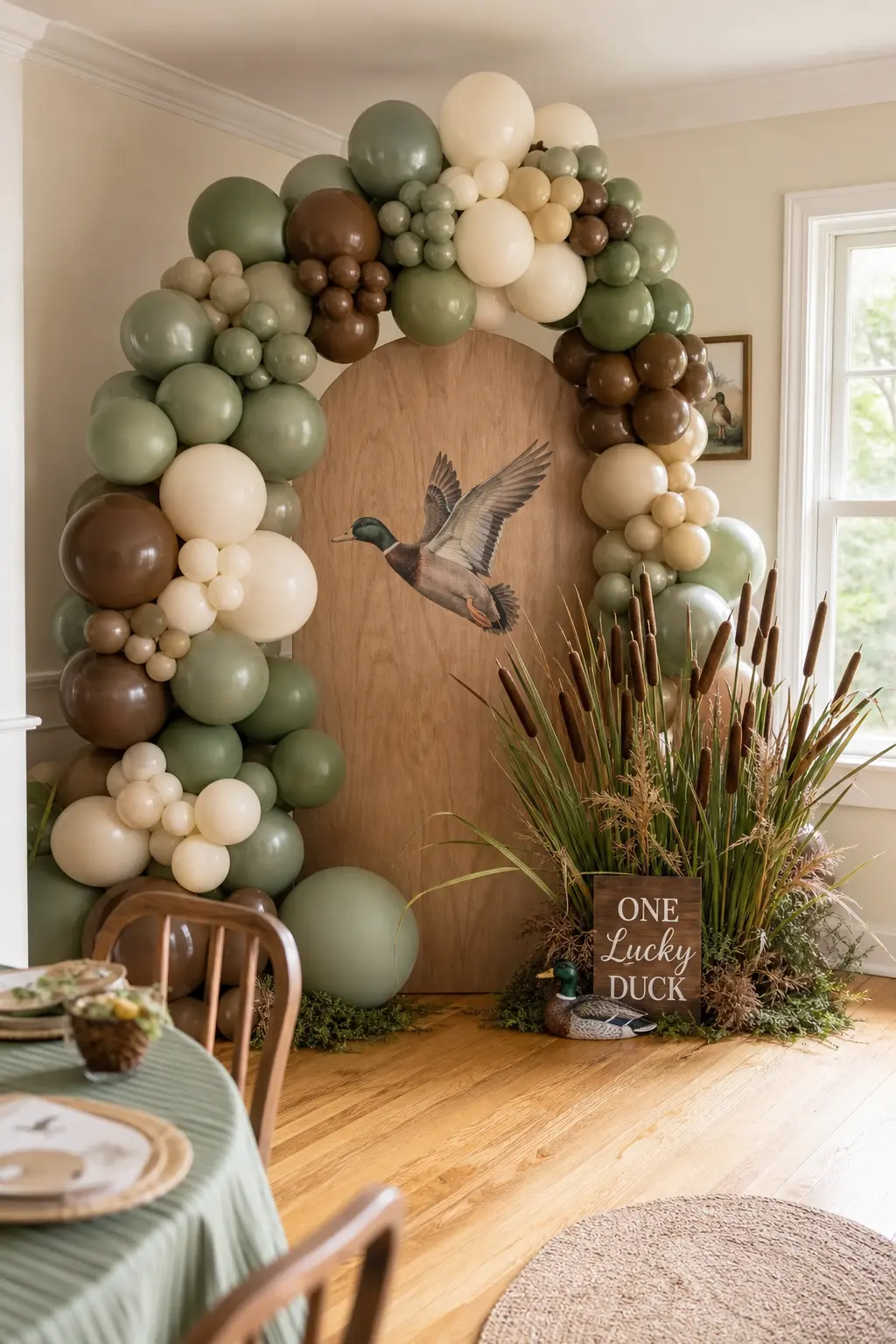 Sage And Walnut Balloon Arch Corner — An arched wood panel printed with a flying mallard becomes the anchor, with an organic balloon garland in sage, olive