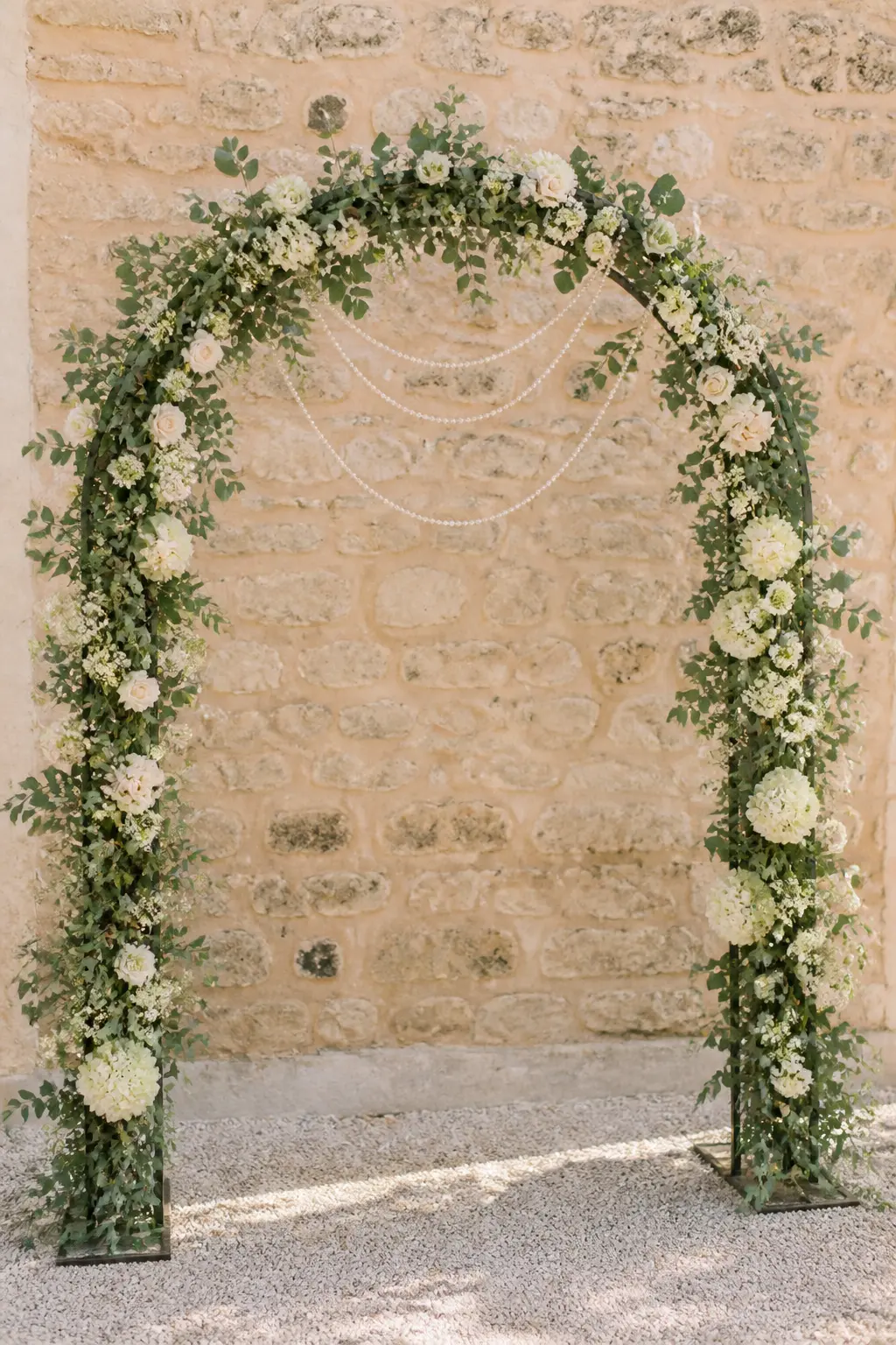 Pearl-Draped Flower Arch — A simple metal arch covered in eucalyptus and white blooms, with pearl garland looped across the top arc in three to five gentle