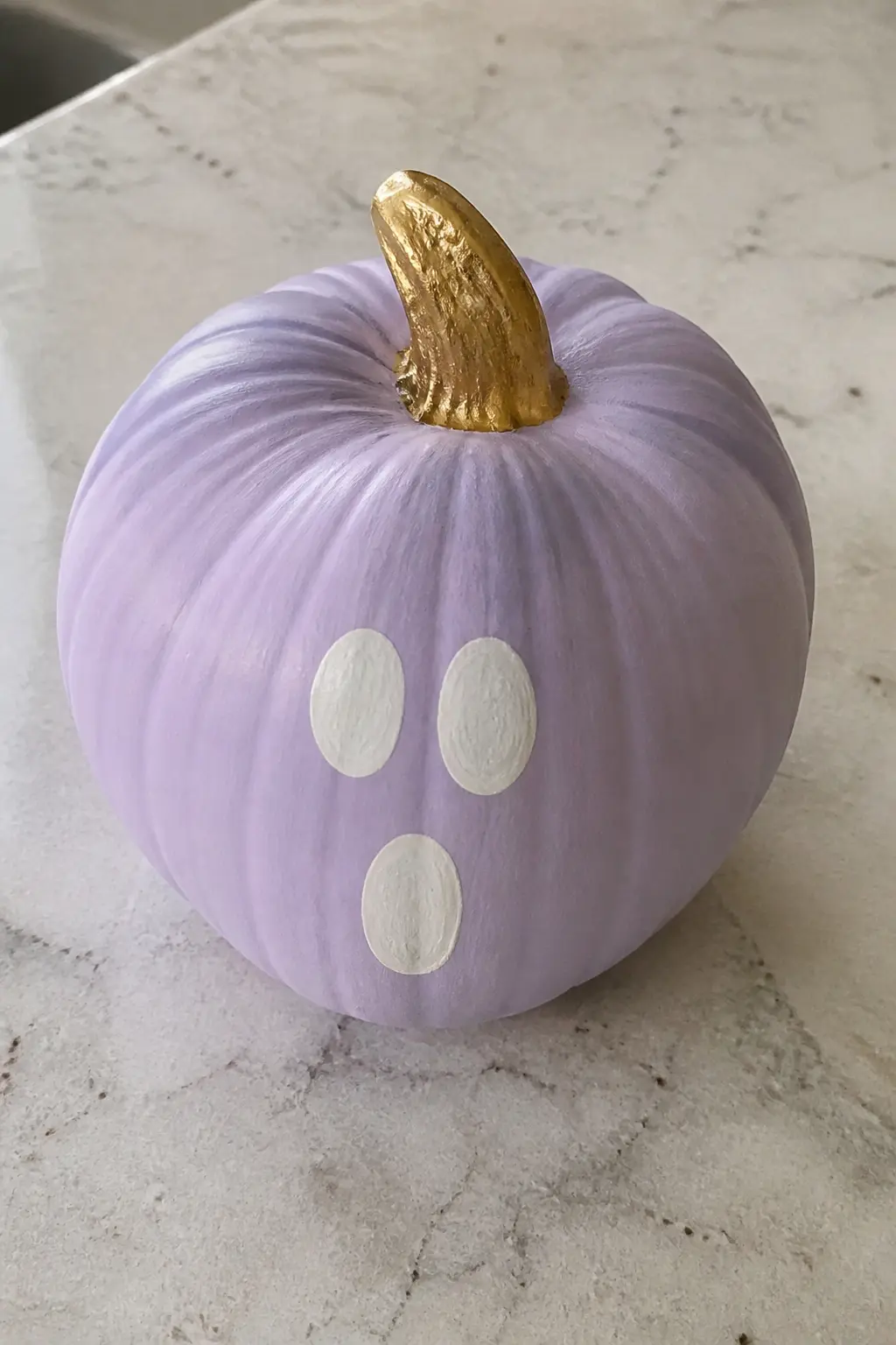 Lavender With a White Ghost Face and a Gold Stem — A lavender base coat already pulls this pumpkin away from standard Halloween orange. Purple painted pumpkin