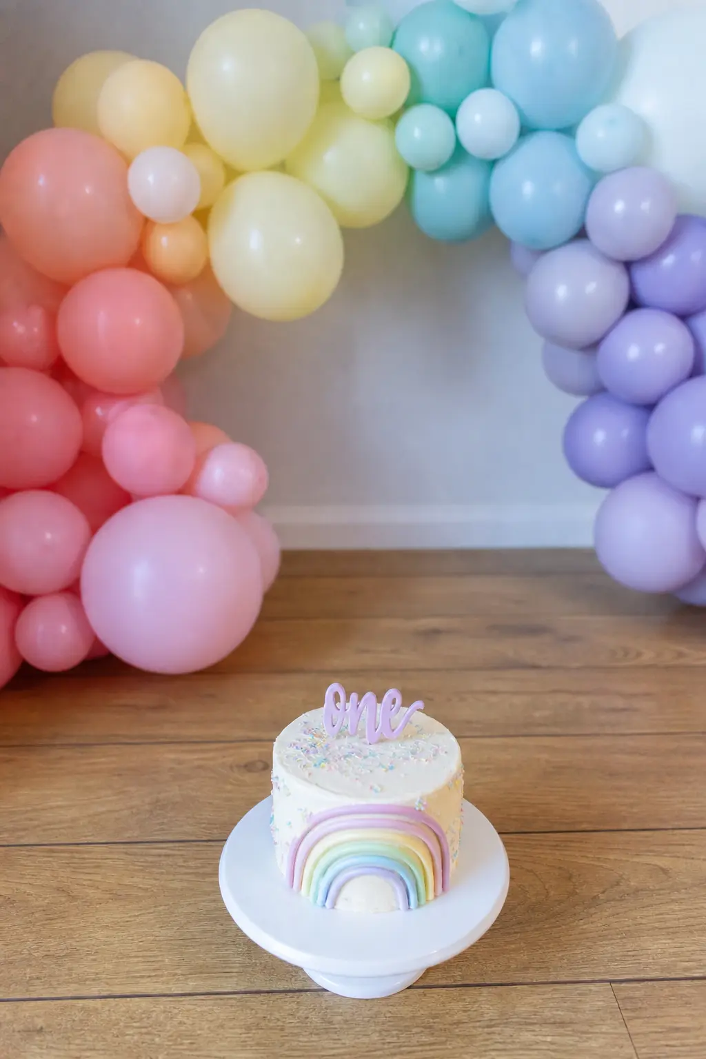 Pastel Spectrum Over a First Birthday Cake — Blush, peach, lemon, mint, periwinkle and lavender in spectrum order, no primary colours anywhere in the arch.