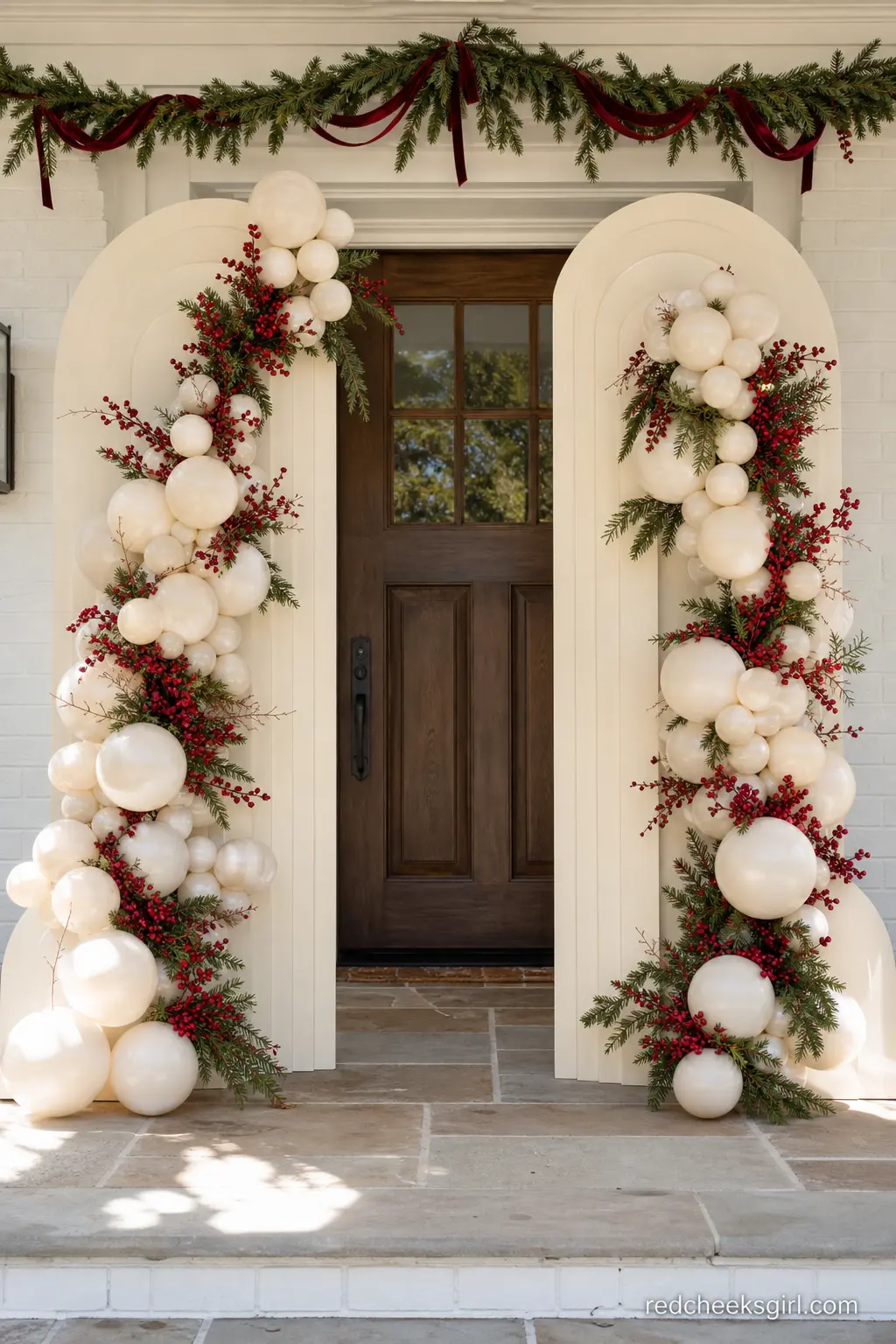 Ivory Split-Arch Backdrop with Cascading Berry Garland — Two cream panel arches flank the front door, each trailing a cascade of ivory balloons, pine sprigs