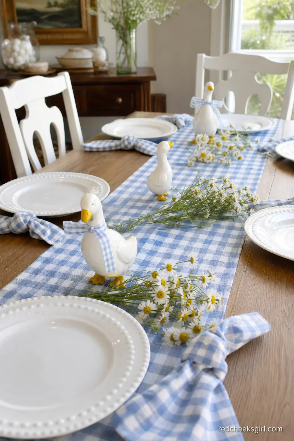 Farmhouse Gingham Table — A long table gets a blue gingham runner down the center, with a trio of small white geese figurines marching along it and loose daisy
