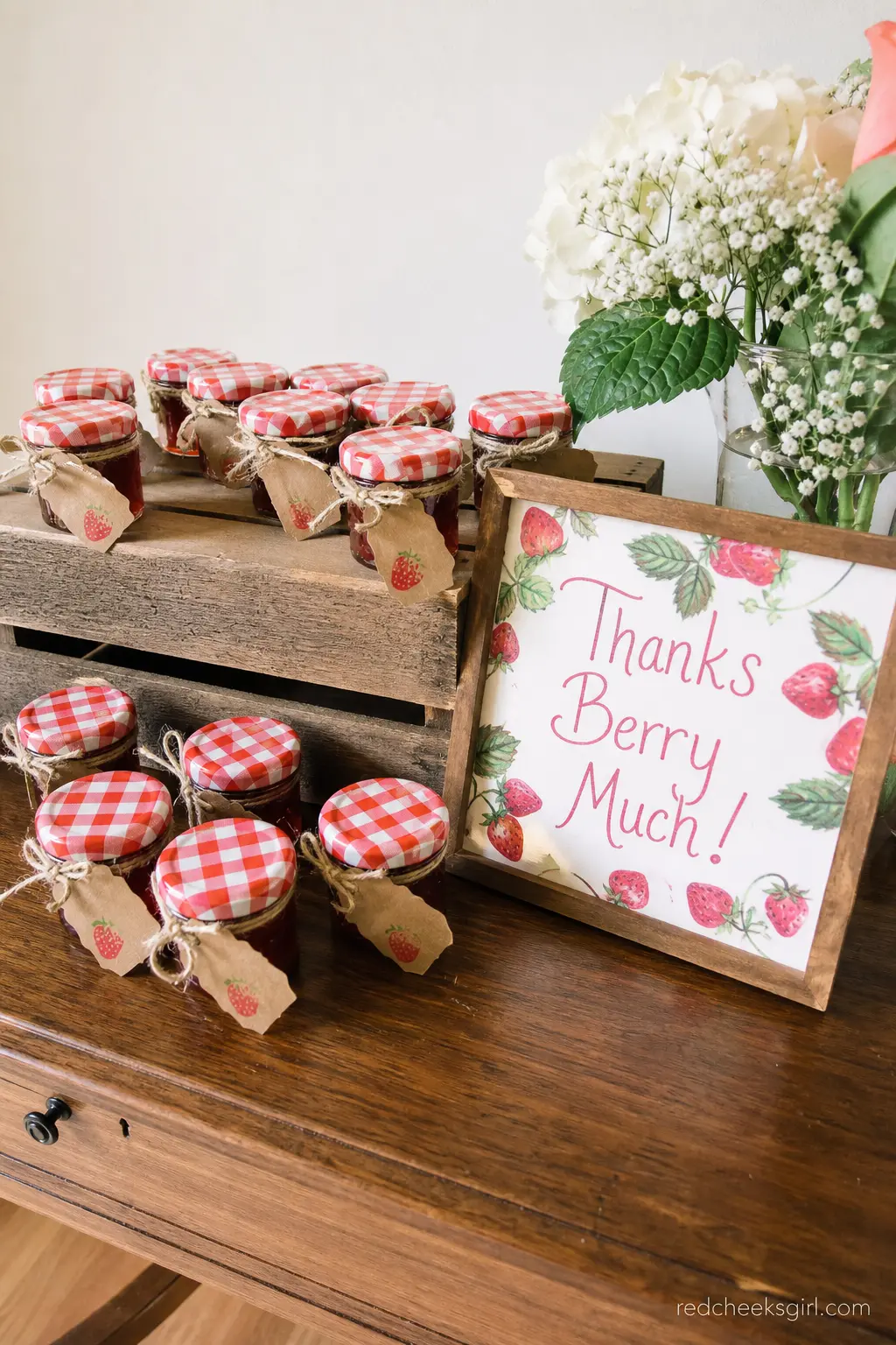 Mini Jam Jar Favor Display — Small jars of strawberry jam with gingham lids and kraft paper labels line up neatly in a wooden crate, doubling as both display