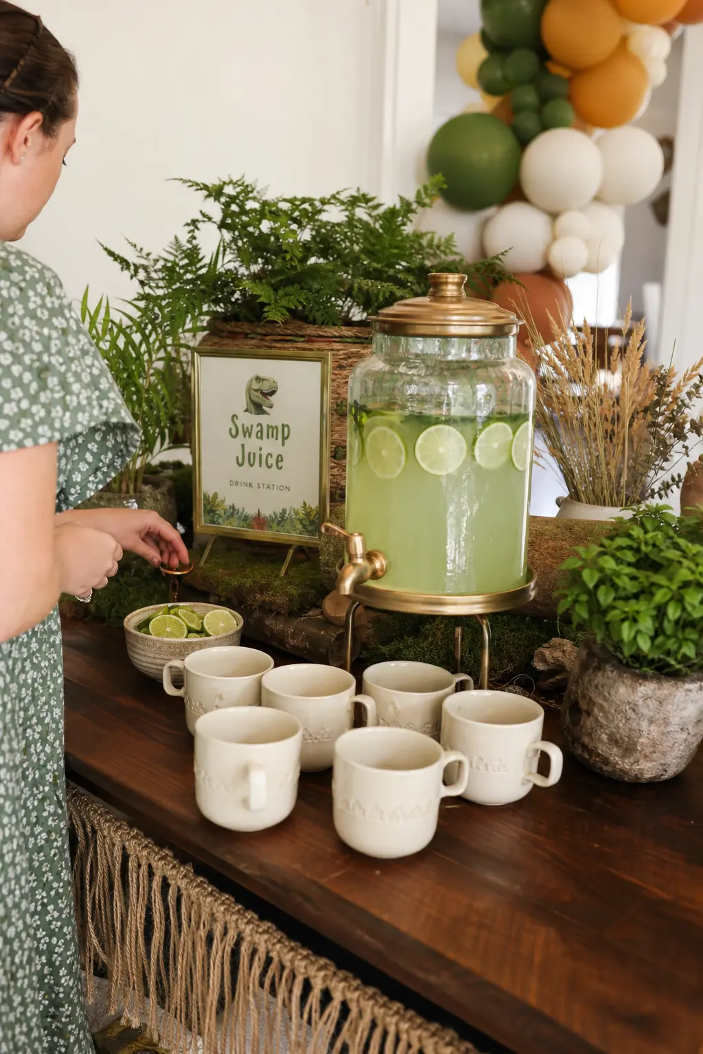 Swamp Juice Drink Station — A brass beverage dispenser filled with pale green lime and mint infused water, set beside a stack of cream mugs and a small bowl of