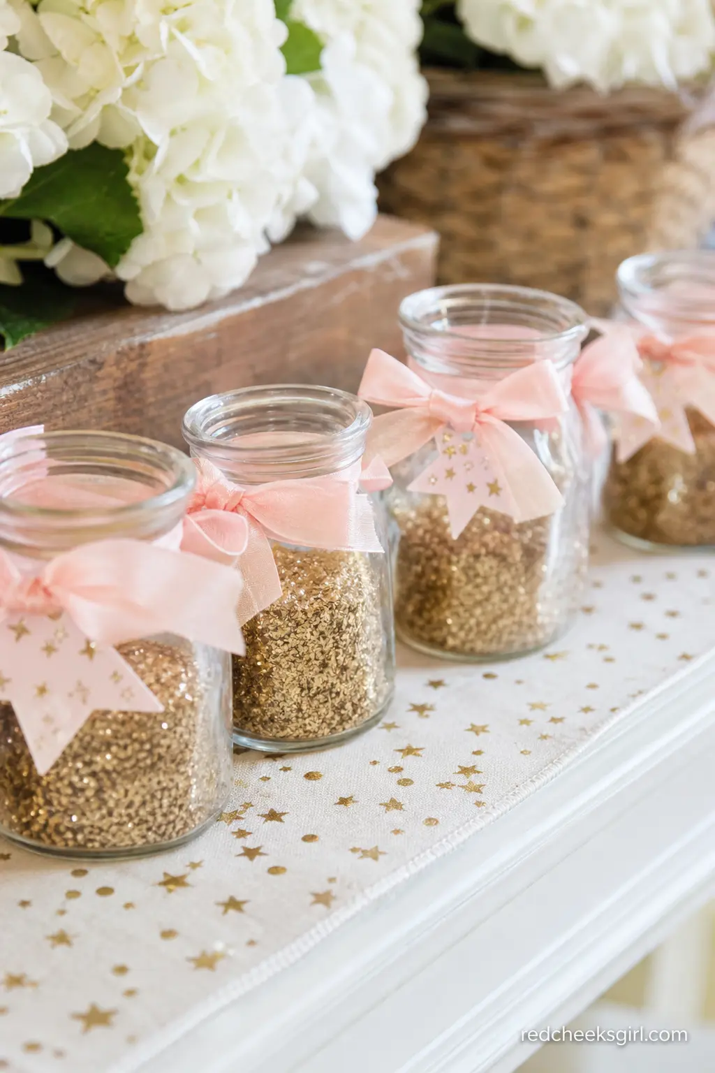 Stardust Favor Jars — Small glass jars filled with fine gold glitter, tied off with a blush ribbon and a tiny star tag, read as stardust the moment guests spot