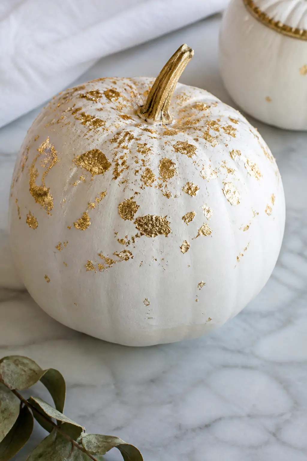 Gold Leaf Flake Scatter — Gold leaf flakes pressed onto a white pumpkin in a loose drift from the top down create something that looks genuinely expensive and