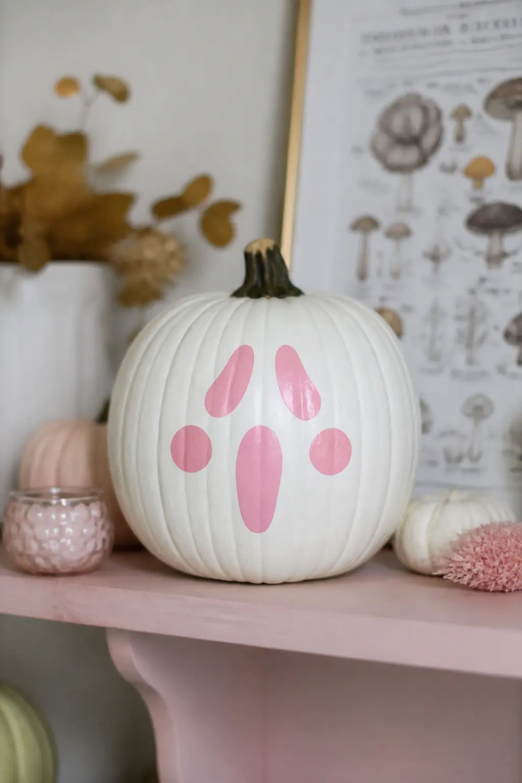 Pink Ghost Face With Rosy Cheeks — A simple ghost face painted in soft pink sits centred on the front of the pumpkin: two round eyes, a small open oval for a