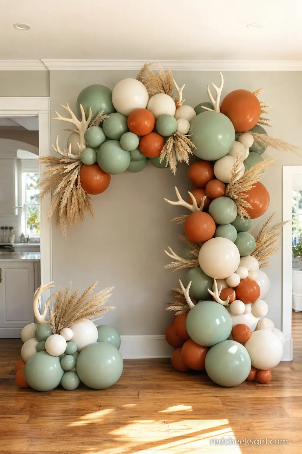 A Balloon and Antler Backdrop in Sage and Rust — An organic balloon garland in sage, rust and cream, threaded with a few faux antlers and dried pampas, gives