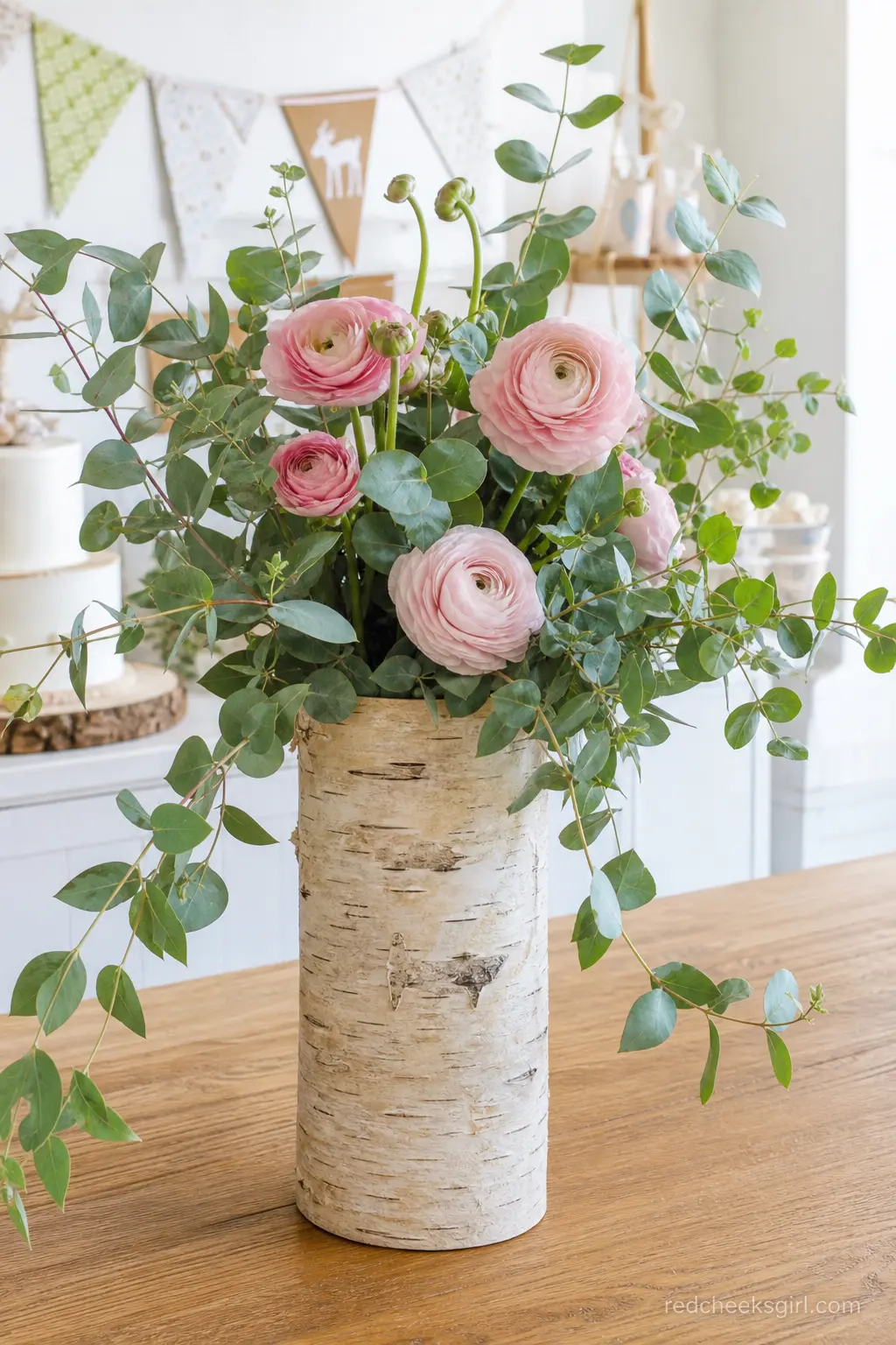 A Birch Bark Vase of Eucalyptus and Ranunculus — A single tall vase wrapped in birch bark paper, filled with trailing eucalyptus and a few dusty pink