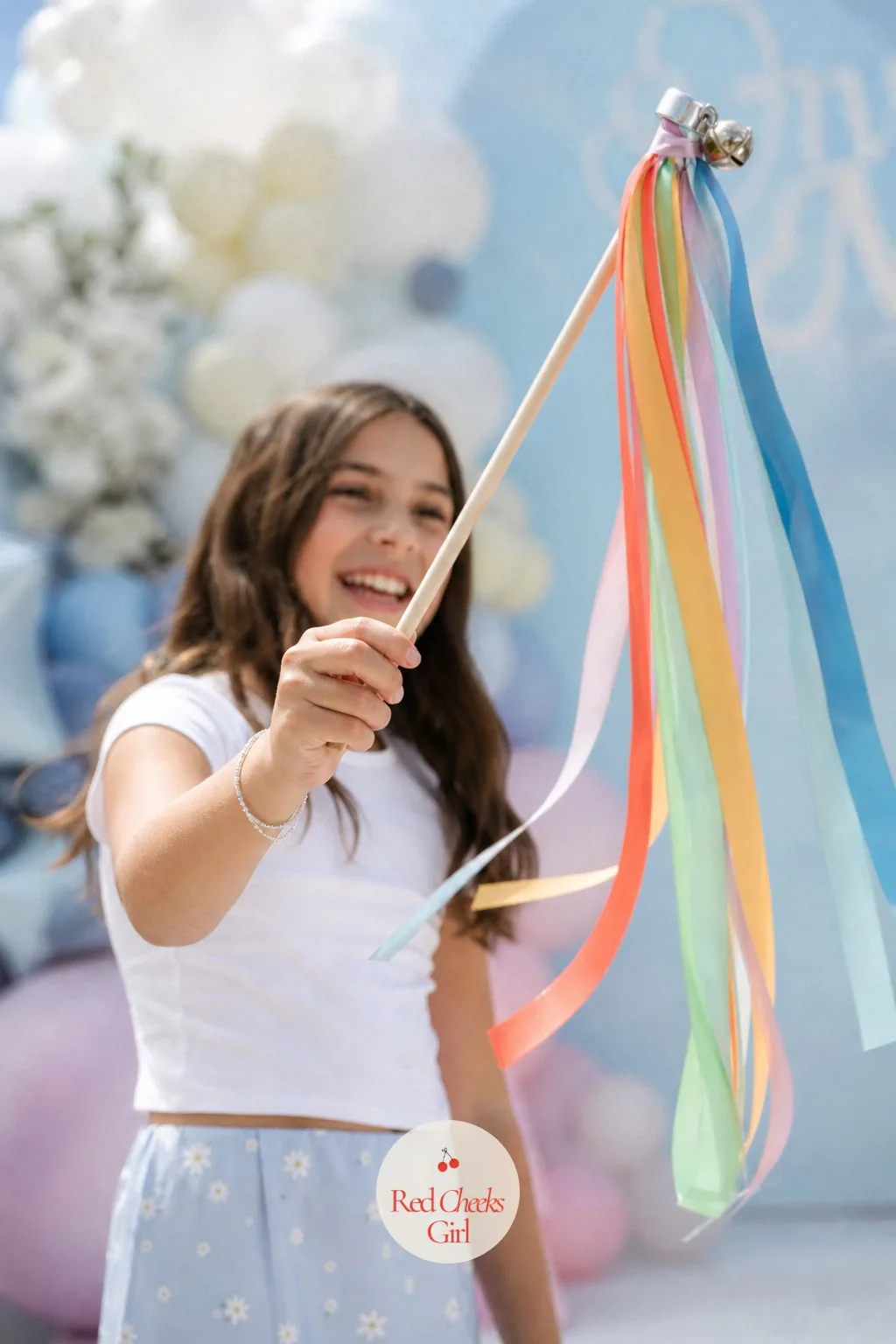 A DIY Ribbon Wand Favor — A wooden dowel or ring forms the handle, with lengths of rainbow and sky blue ribbon tied on in a cluster so they flutter loose at