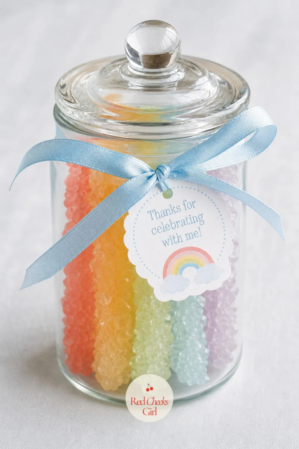 A Mini Rainbow Rock Candy Jar — A small glass jar holds a handful of rainbow rock candy sticks standing upright inside, the lid tied off with a length of sky