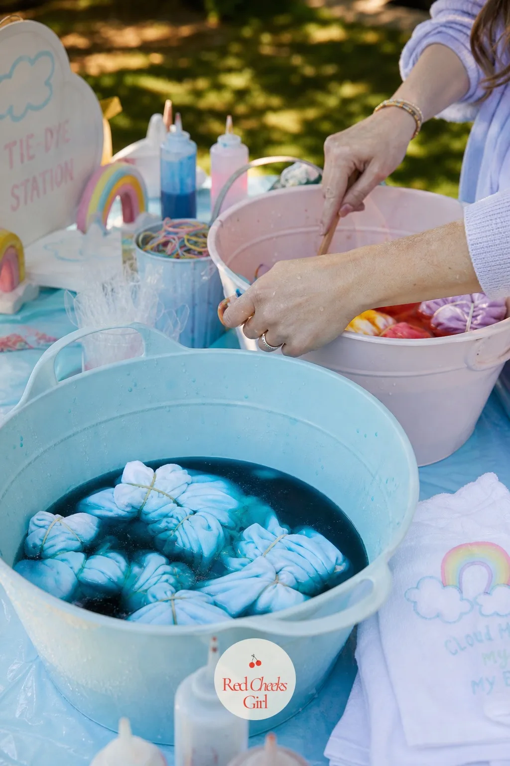 A Tie-Dye Station — A plastic sheet covers the table first, then plain white cotton shirts or tote bags sit soaking in buckets of sky blue and rainbow dye