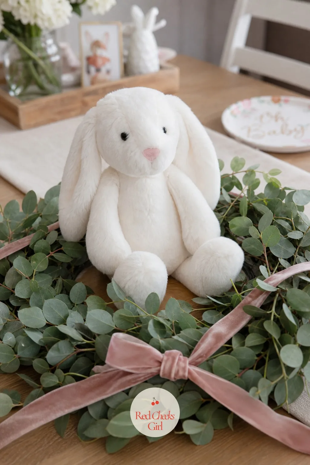Bunny Plush With a Wreath — A white bunny plush sits at the center of a small eucalyptus wreath laid flat on the table, dusty pink ribbon crossing the top of