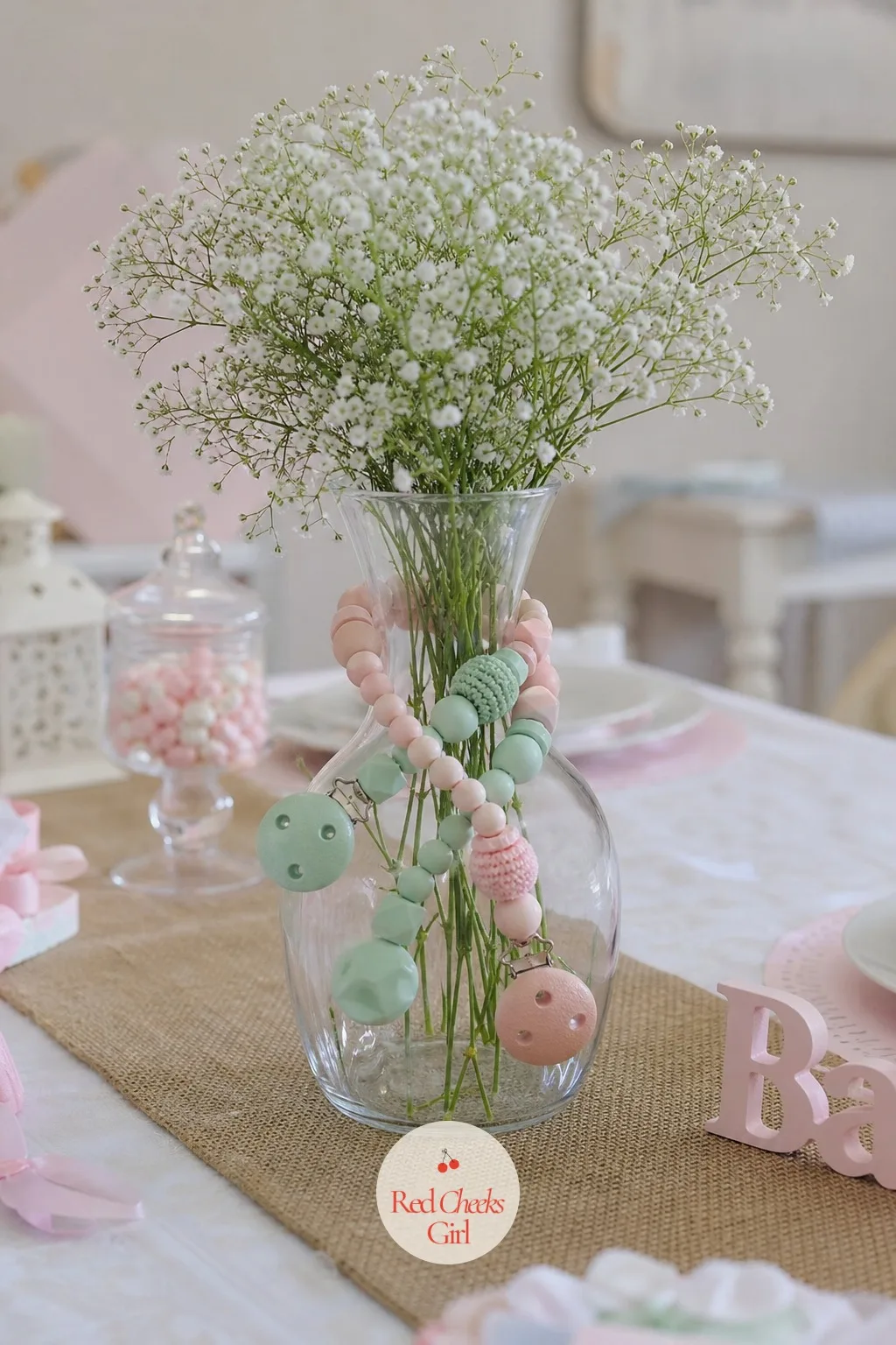 Pacifier Clips Around a Vase — A dozen pacifier clips, ribbon ends dangling, loop around the neck of a clear vase like a collar, pastel pink and mint