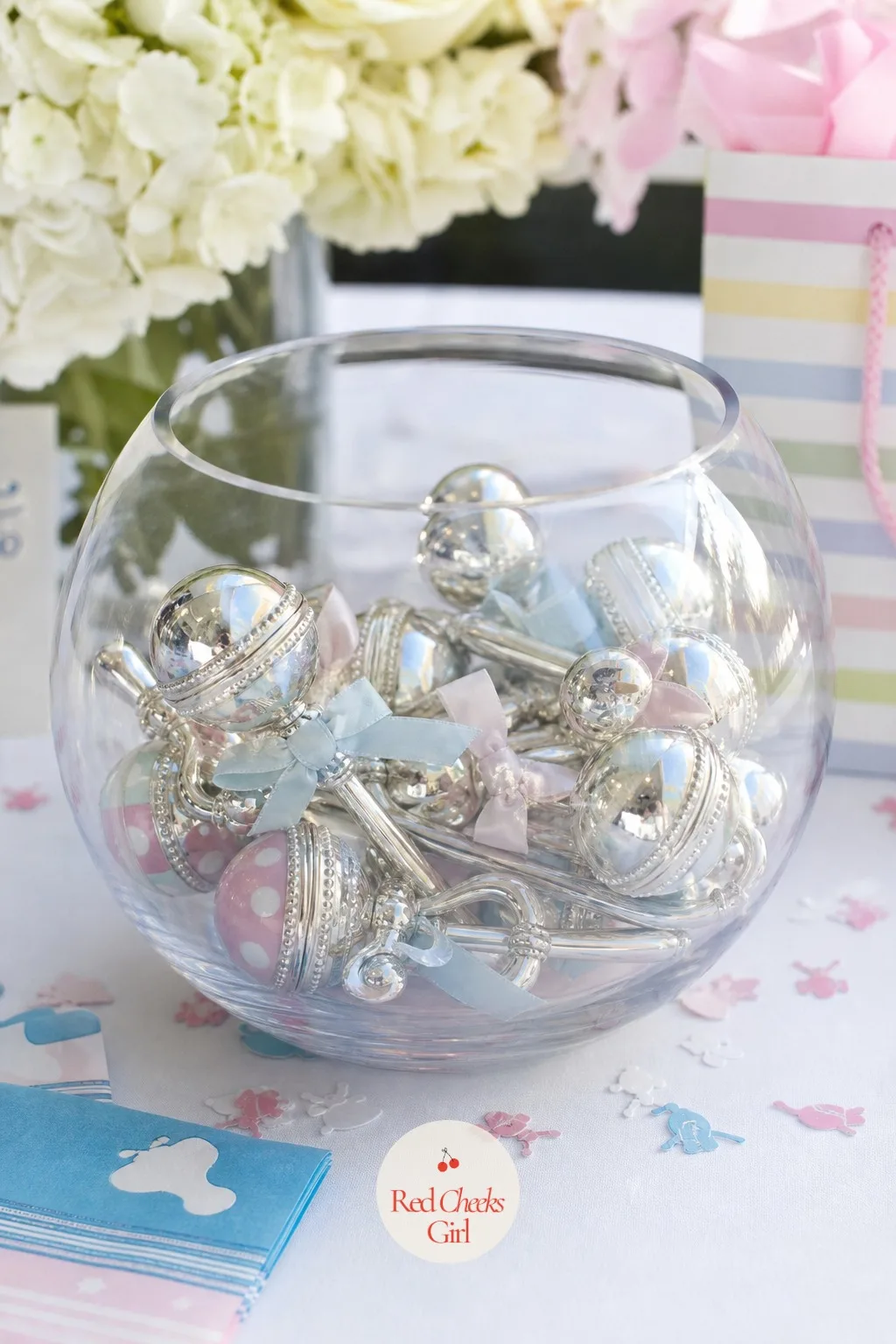 Rattles in a Glass Bowl — Silver and pastel rattles, the kind that come individually wrapped in most registries, pile loosely into a clear glass bowl the way