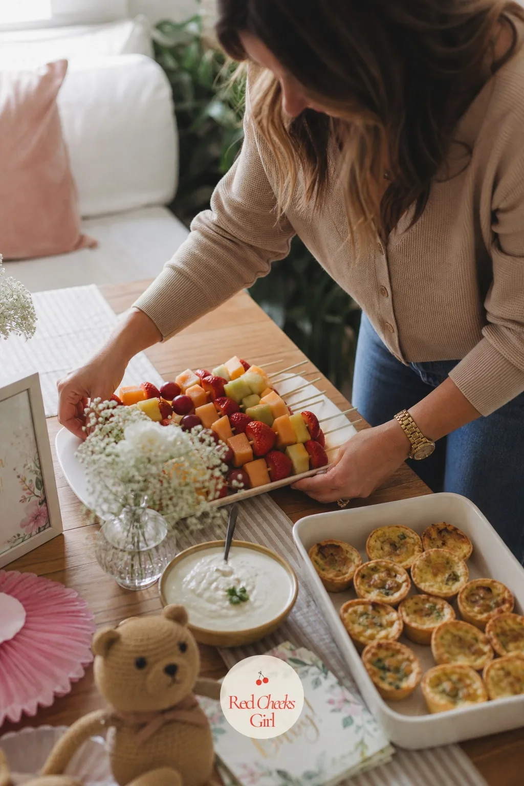 Plan the Menu Around What She Can Actually Eat — A food table holds small platters mid-assembly: fruit skewers, a bowl of dip, a tray of mini quiches still