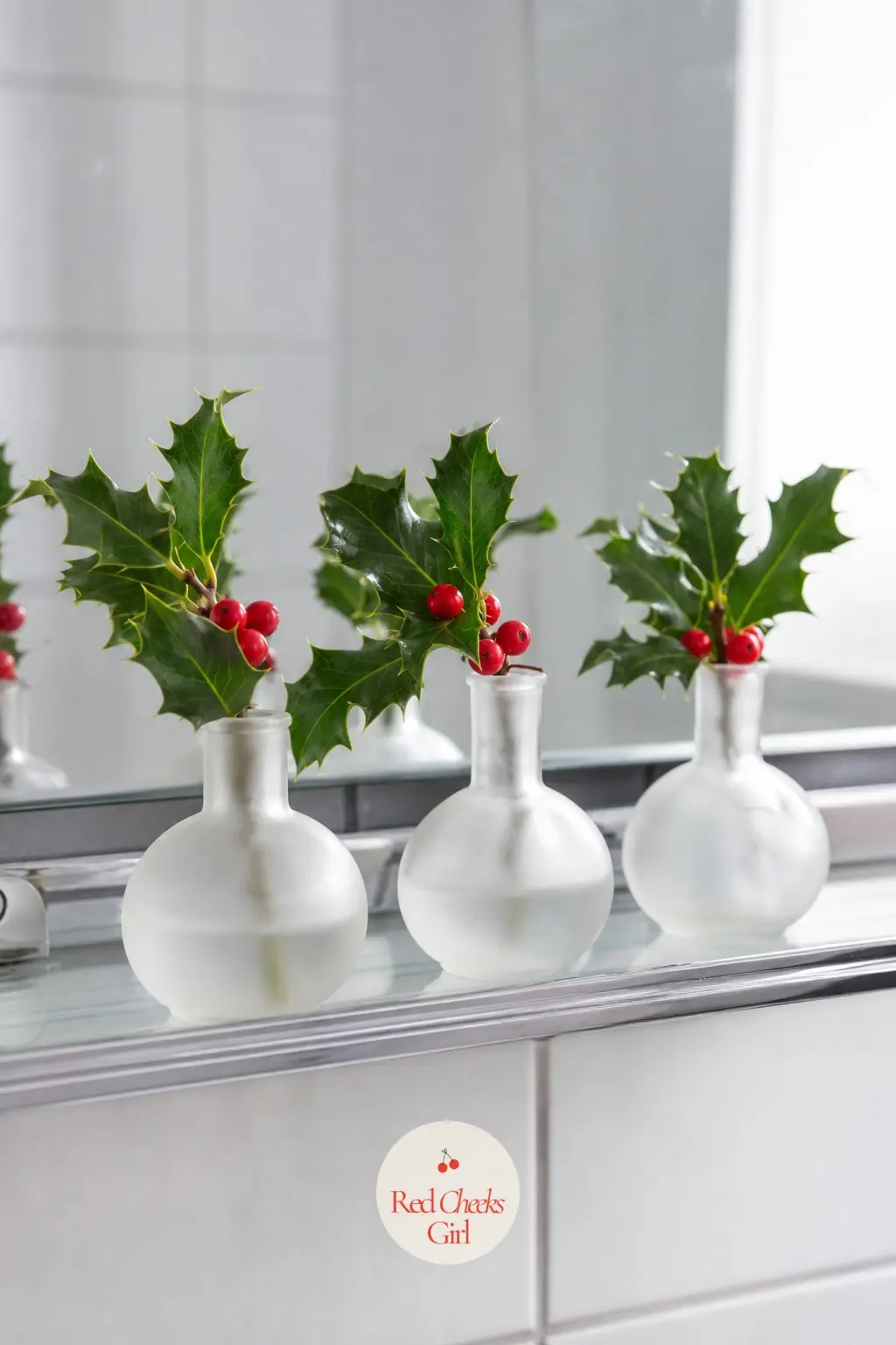 A Trio of Frosted Glass Bud Vases Lined Along the Mirror Ledge, Each Holding One Sprig of Holly — Three identical frosted glass bud vases, lined up in a row