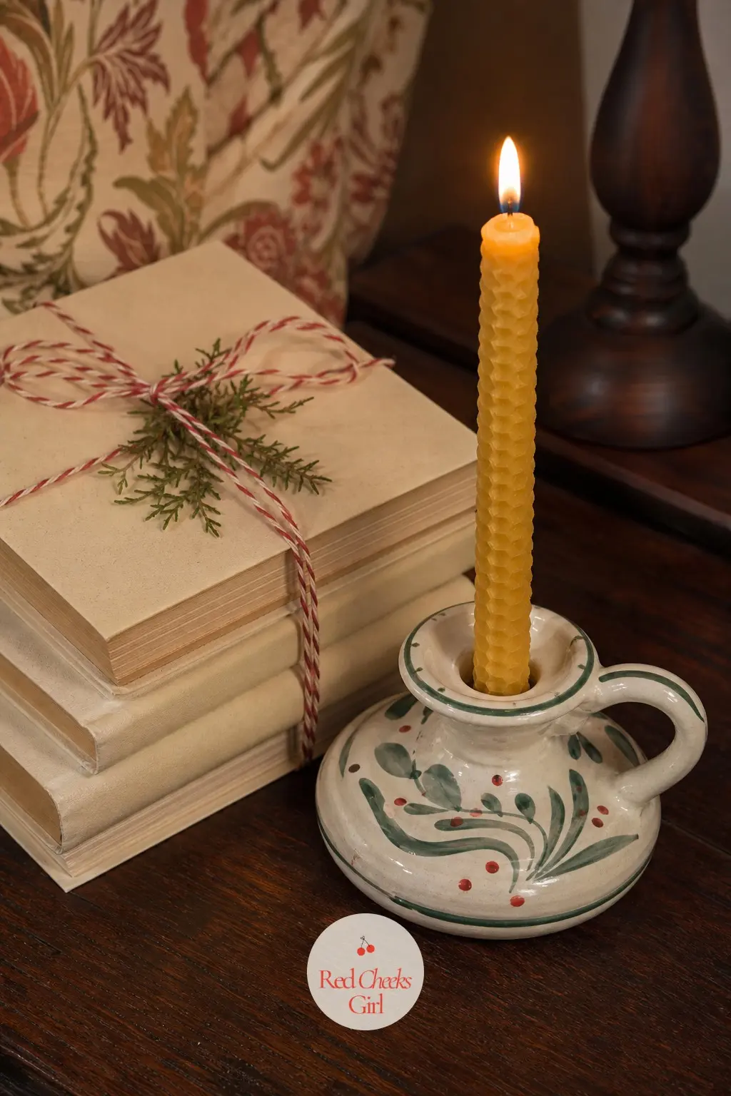 A Beeswax Taper in a Painted Ceramic Holder Beside Wrapped Books — One lit beeswax taper, set in a small hand painted ceramic holder, next to two or three