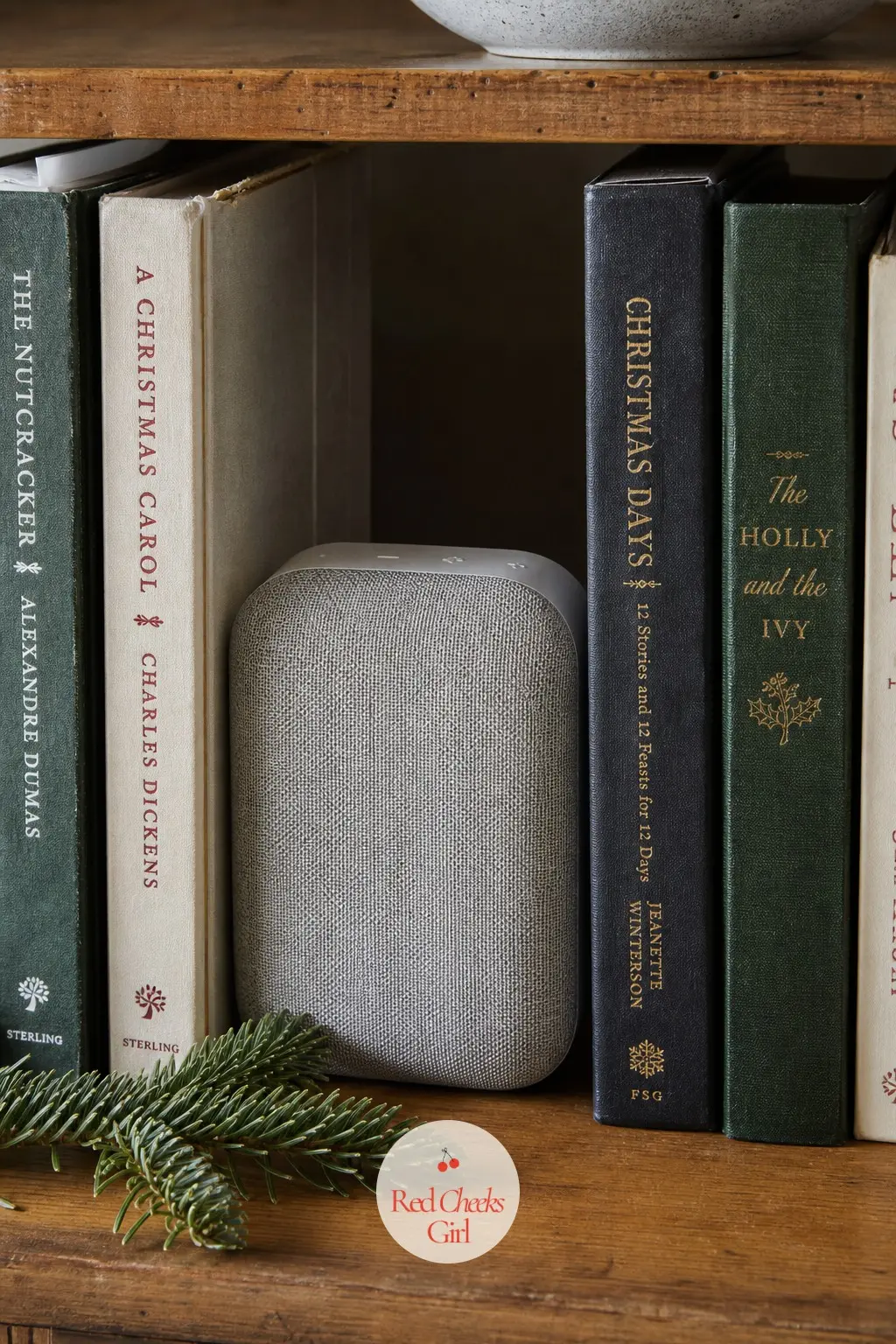 A Small Speaker Tucked Among the Books Playing Soft Carols — A compact speaker, fabric front in a soft heather grey, sits on a shelf among the books rather