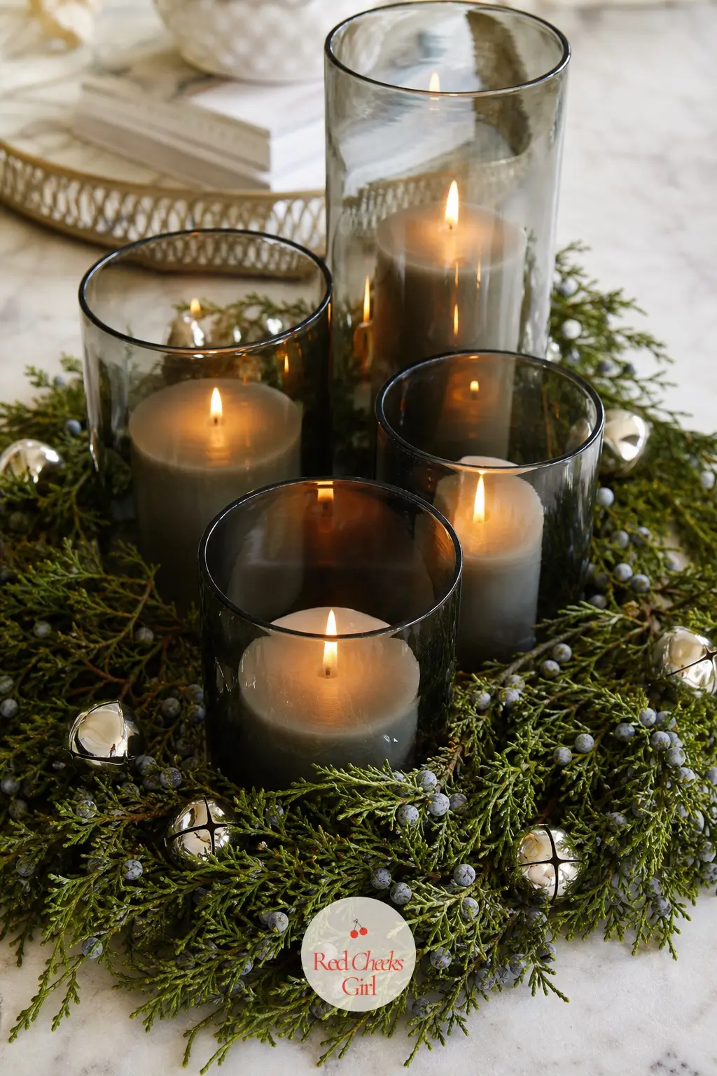 A Ring of Smoked Glass Pillars Circled With Juniper and Silver Bells — A cluster of smoked grey glass pillar candles sits ringed by sprigs of fresh juniper