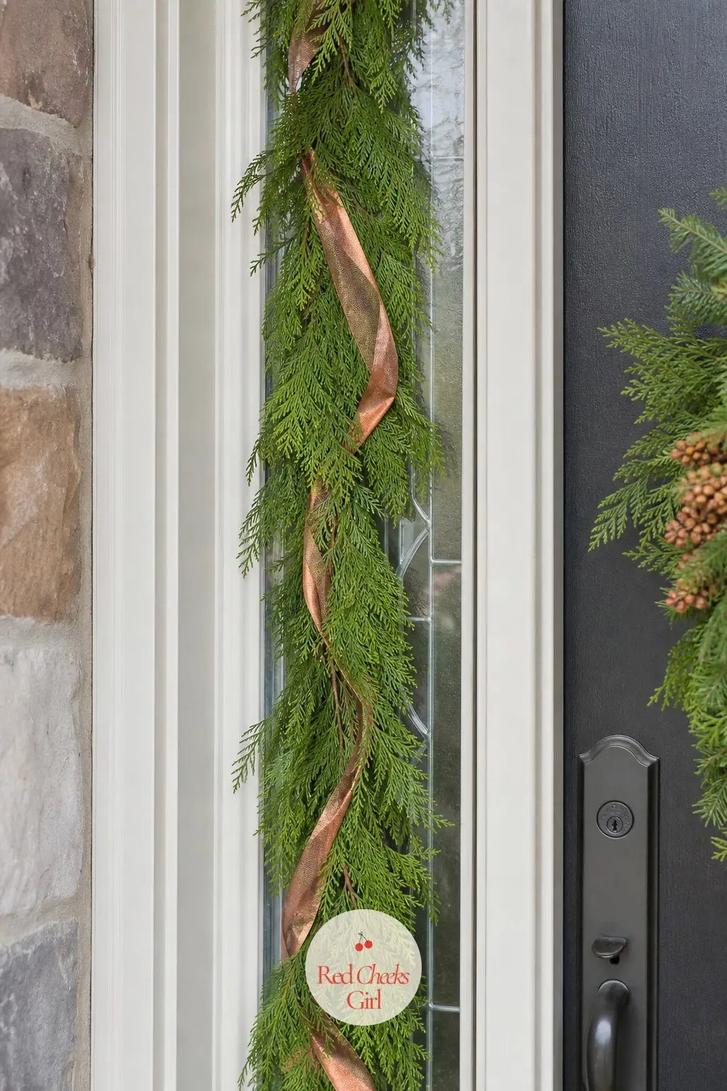 A Cedar Garland Threaded Down Both Sidelights — Narrow sidelight windows either side of the door take a thin cedar garland each, copper ribbon woven through at
