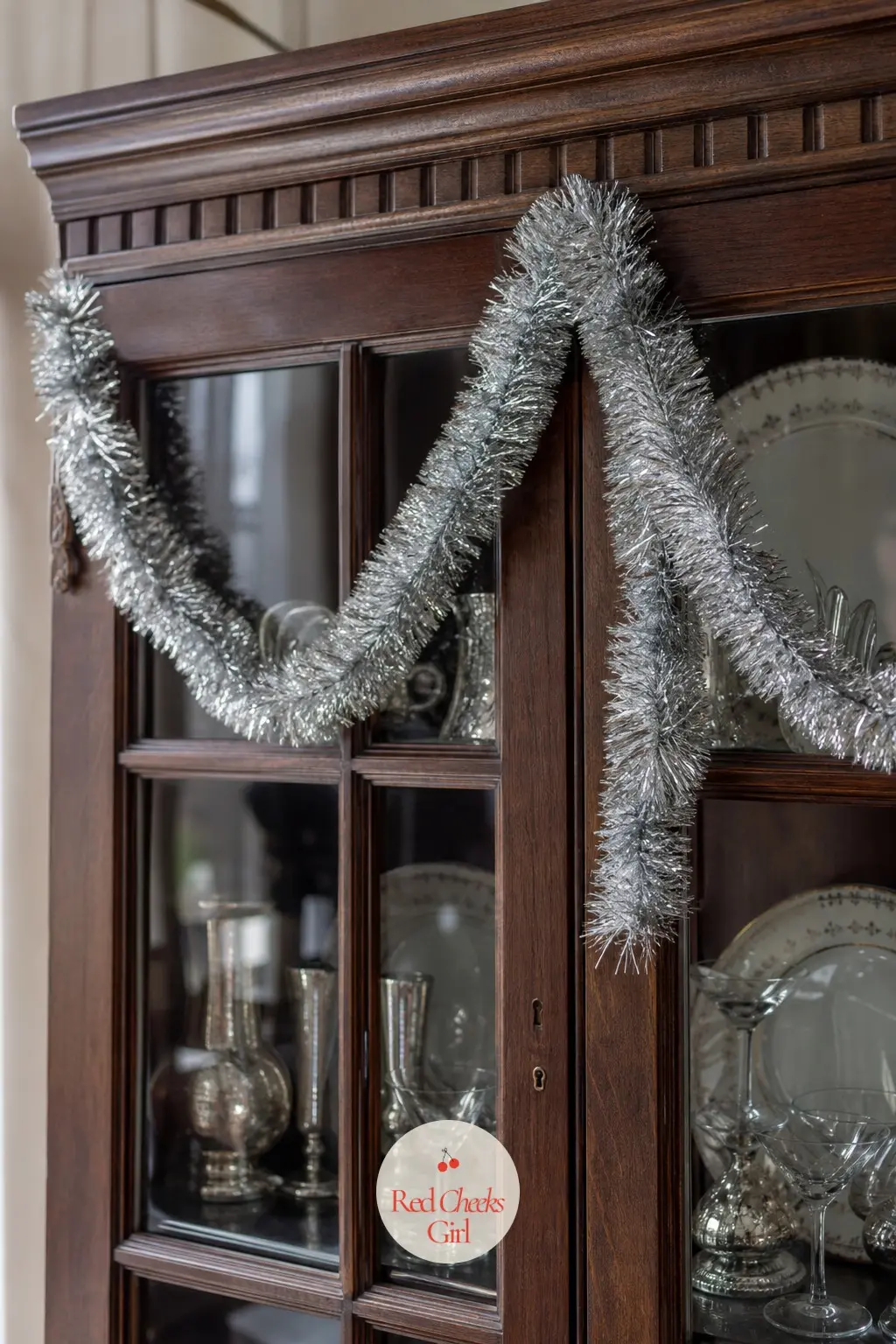Vintage Tinsel Draped In Swags — Silver tinsel draped in loose swags across a china cabinet front is unapologetically retro, the kind of garland a