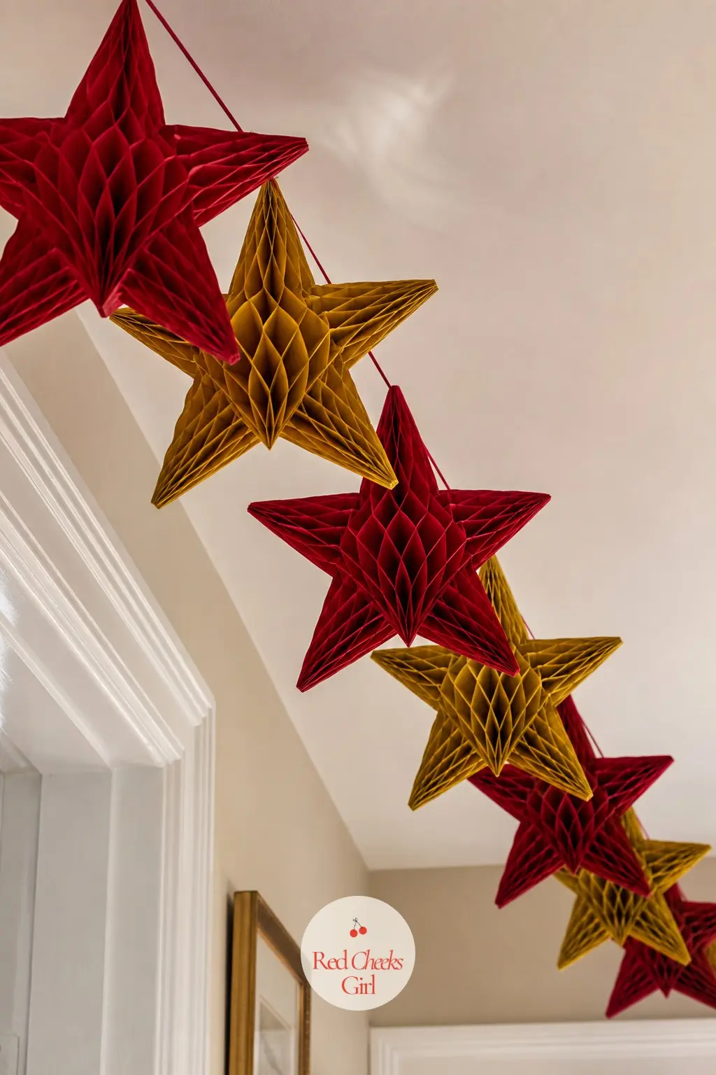 A Honeycomb Paper Star Garland Strung Corner to Corner Across the Ceiling — Tissue honeycomb stars in deep red and gold, strung on a single cord corner to