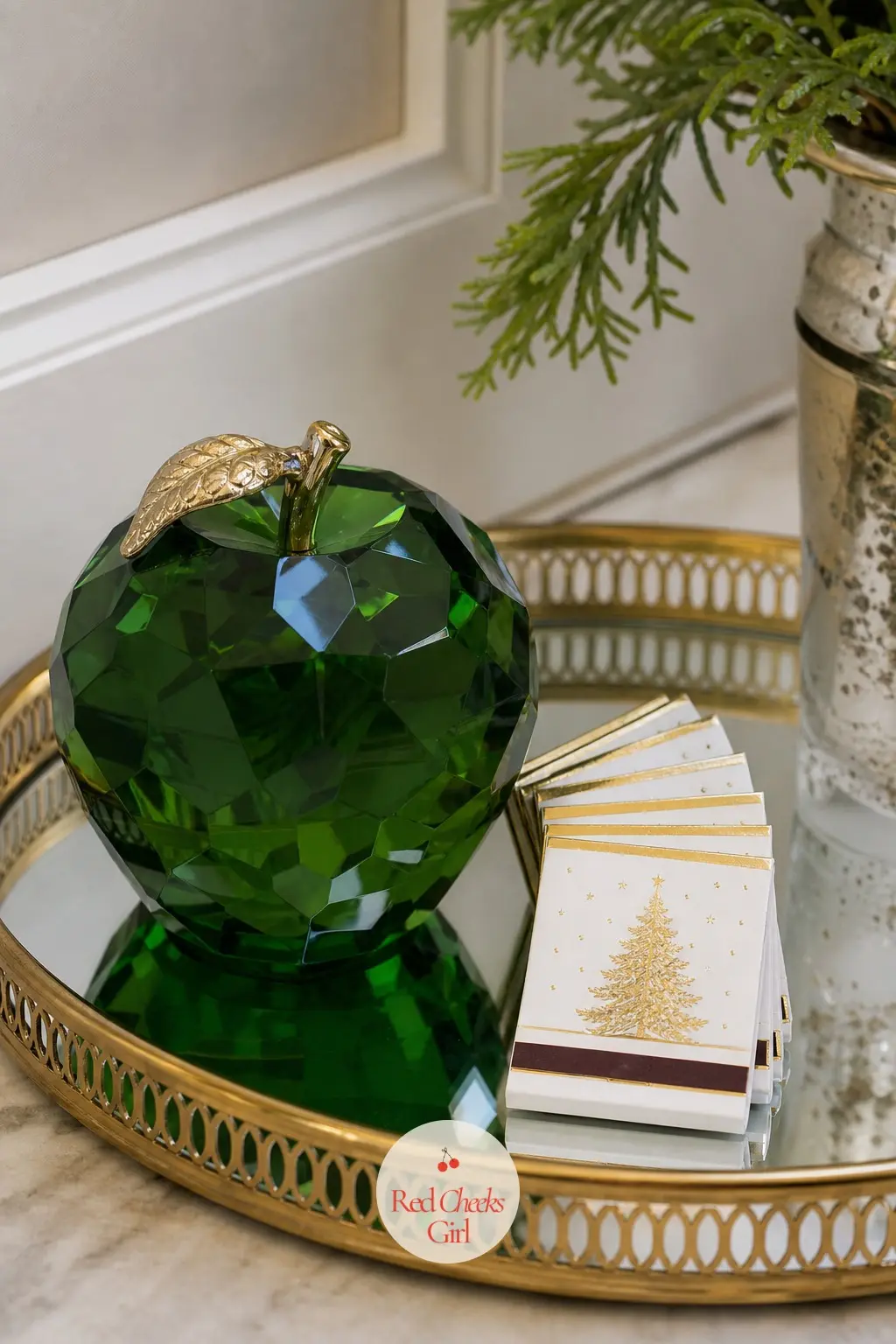 A Mirrored Tray With a Faceted Glass Apple and Gold Rimmed Matchbooks — A single faceted glass apple in deep emerald sits at the centre of a mirrored tray