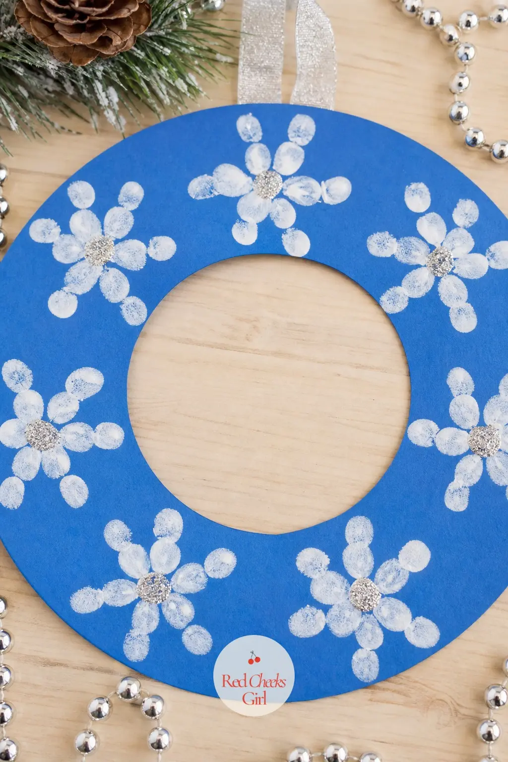 Thumbprint Snowflake Wreath — On a blue cardstock ring, cluster white paint thumbprints into six-point snowflake shapes spaced evenly around the circle.