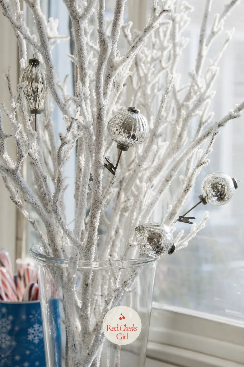 A Frosted Branch Arrangement in a Glass Vase on the Windowsill — Stand a bundle of white flocked branches in a clear glass vase on the windowsill and clip a