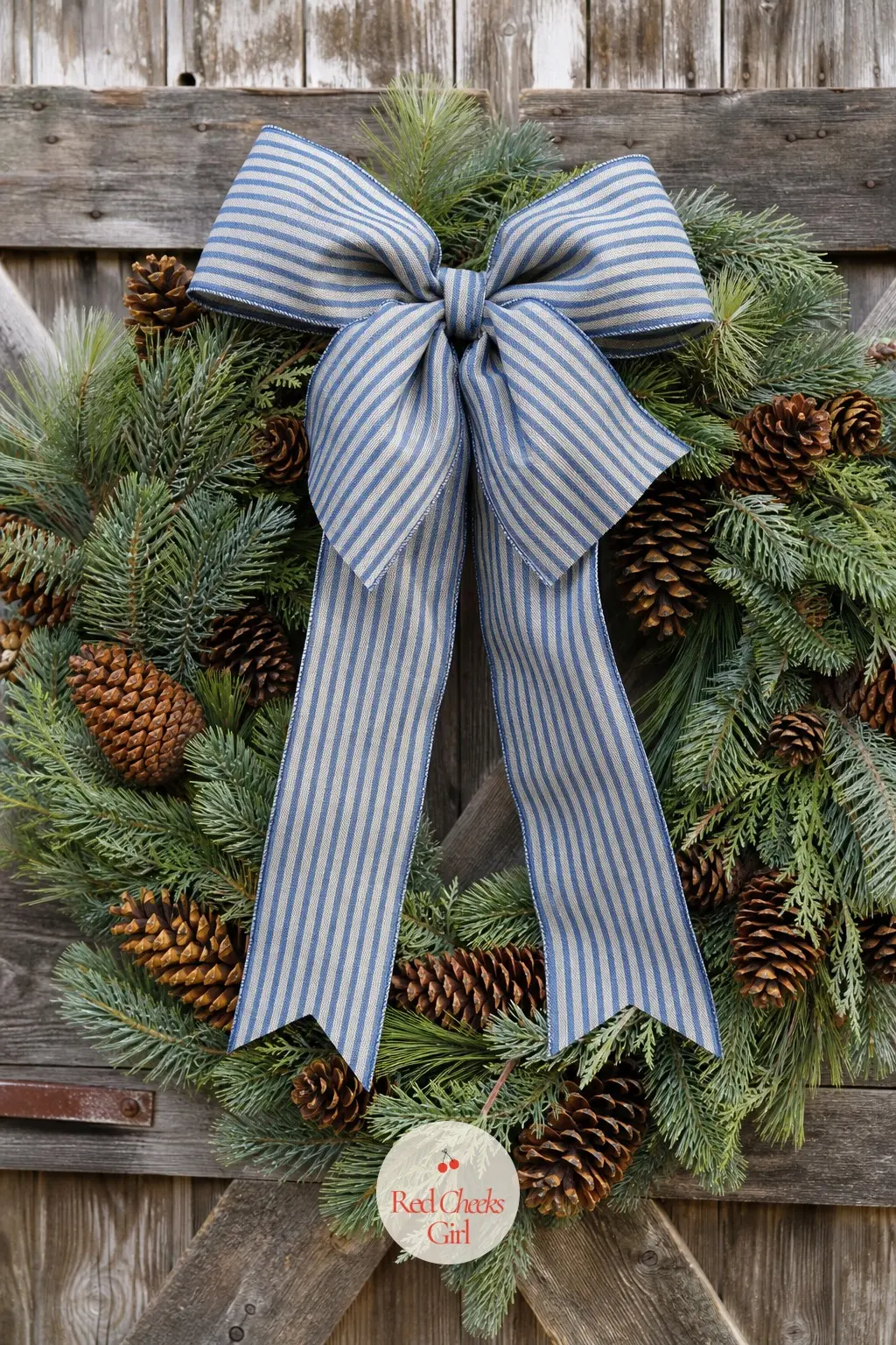 The Ticking Stripe Bow and Pine Cone Wreath for the Barn Door — A denim-blue ticking stripe bow anchors a loose ring of grey-green pine and natural pinecones