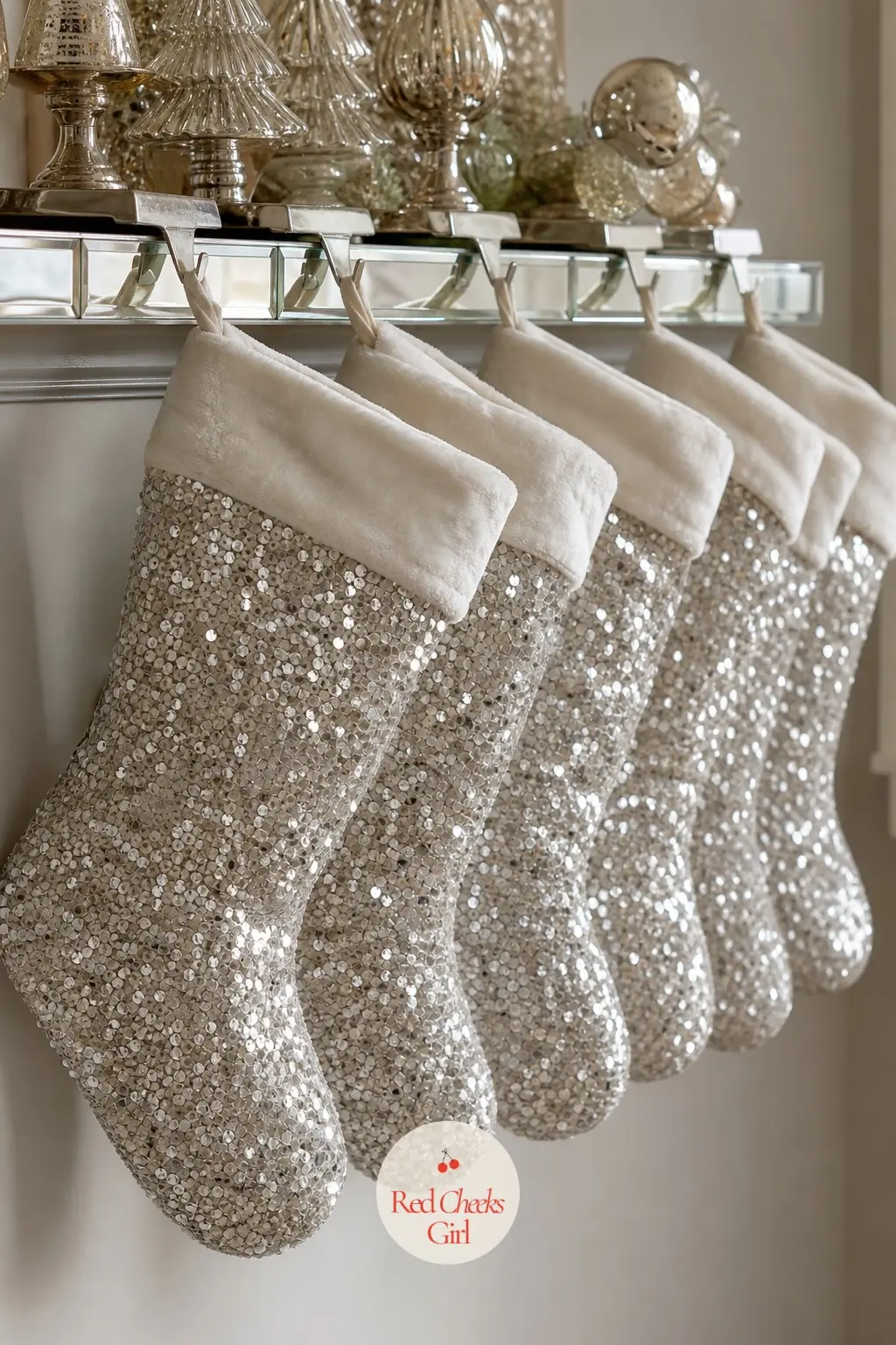 The Stocking Wall, Hung in Sequin and Silver — Six sequin stockings hang from a mirrored hook rail rather than a mantel edge, spaced evenly so each one catches