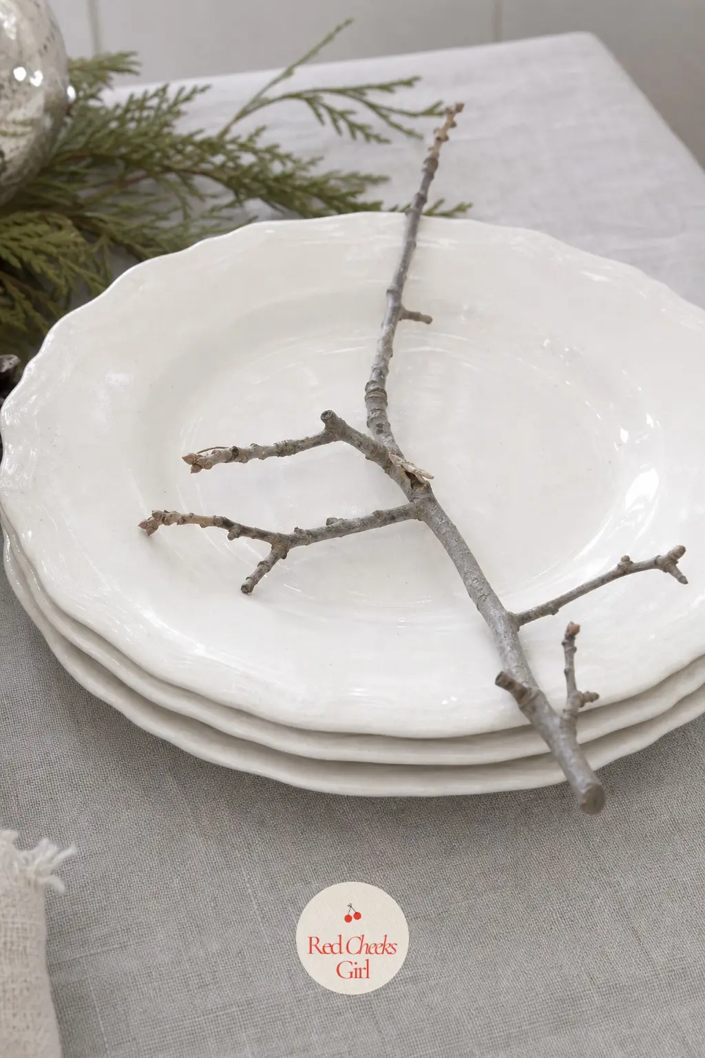 A Stack of Ironstone Plates Topped with a Single Branch — Three white ironstone plates, stacked and set slightly off-center at the head of the table, with one