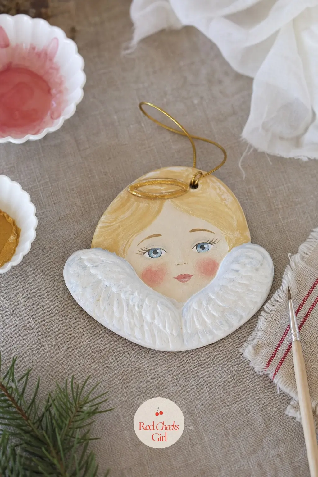 Little Angel Face Ornament — A thin ring of gold paint arcs above the forehead for a halo, floating just clear of the dough rather than touching it, and a soft