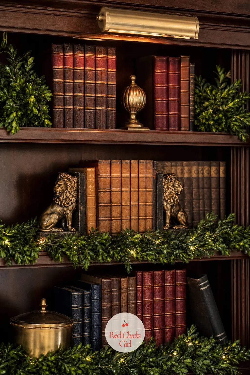 Bookshelves Dressed For The Season — A few stems of faux boxwood tuck along the shelf edges, brass lion bookends hold up a run of leather spines, and one small