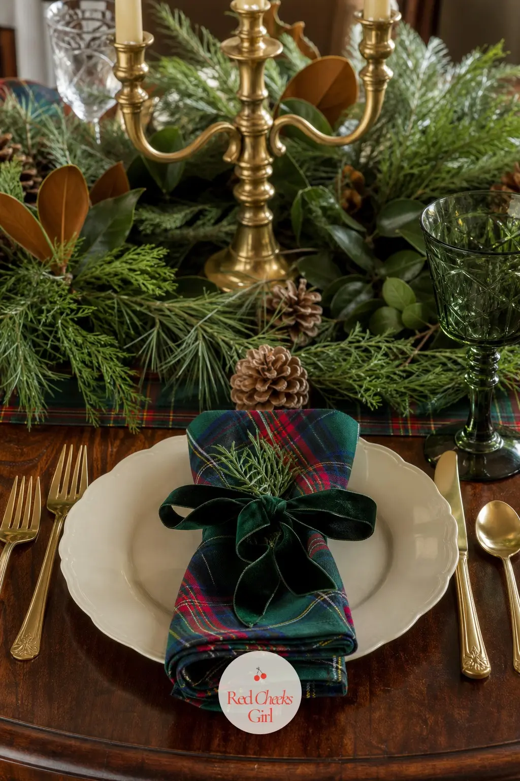 A Centerpiece Of Greenery And One Set Place — A low arrangement of fresh greenery and a few oxblood ranunculus runs between a pair of brass candelabras, and